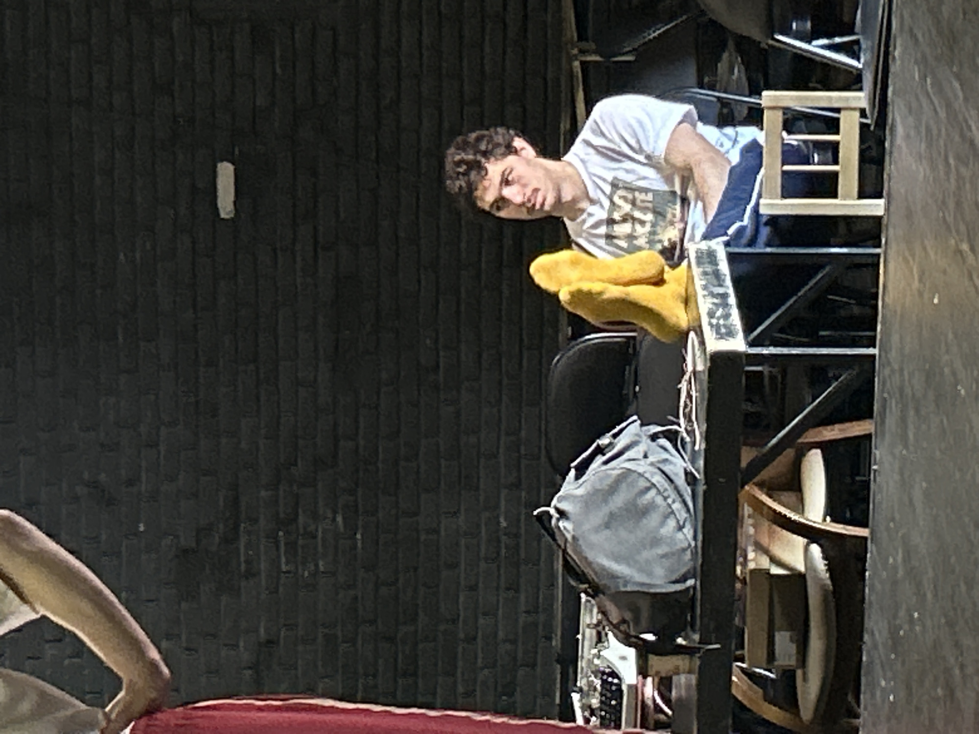 The media file is a vertical, slightly blurry photograph of a young man with curly brown hair wearing a white t-shirt and dark blue shorts with a white stripe. He is looking down and to his left with a curious or concerned expression. His right arm is raised, and he is holding two bright yellow objects that appear to be oven mitts or large gloves. He is positioned on some sort of structure with dark metal supports and wooden elements, resembling scaffolding or stage equipment. To his left, a dark, textured wall is visible, possibly brick. In the foreground, a portion of a red surface, possibly fabric, is seen. To the right of the man, a gray backpack is attached to the structure, and further to the right, more of the metal and wooden framework is visible, along with some rounded, light-colored objects. The lighting suggests it might be daytime, with a light source coming from the top right. The overall scene appears to be backstage or a workshop area. No text is clearly legible in the image.