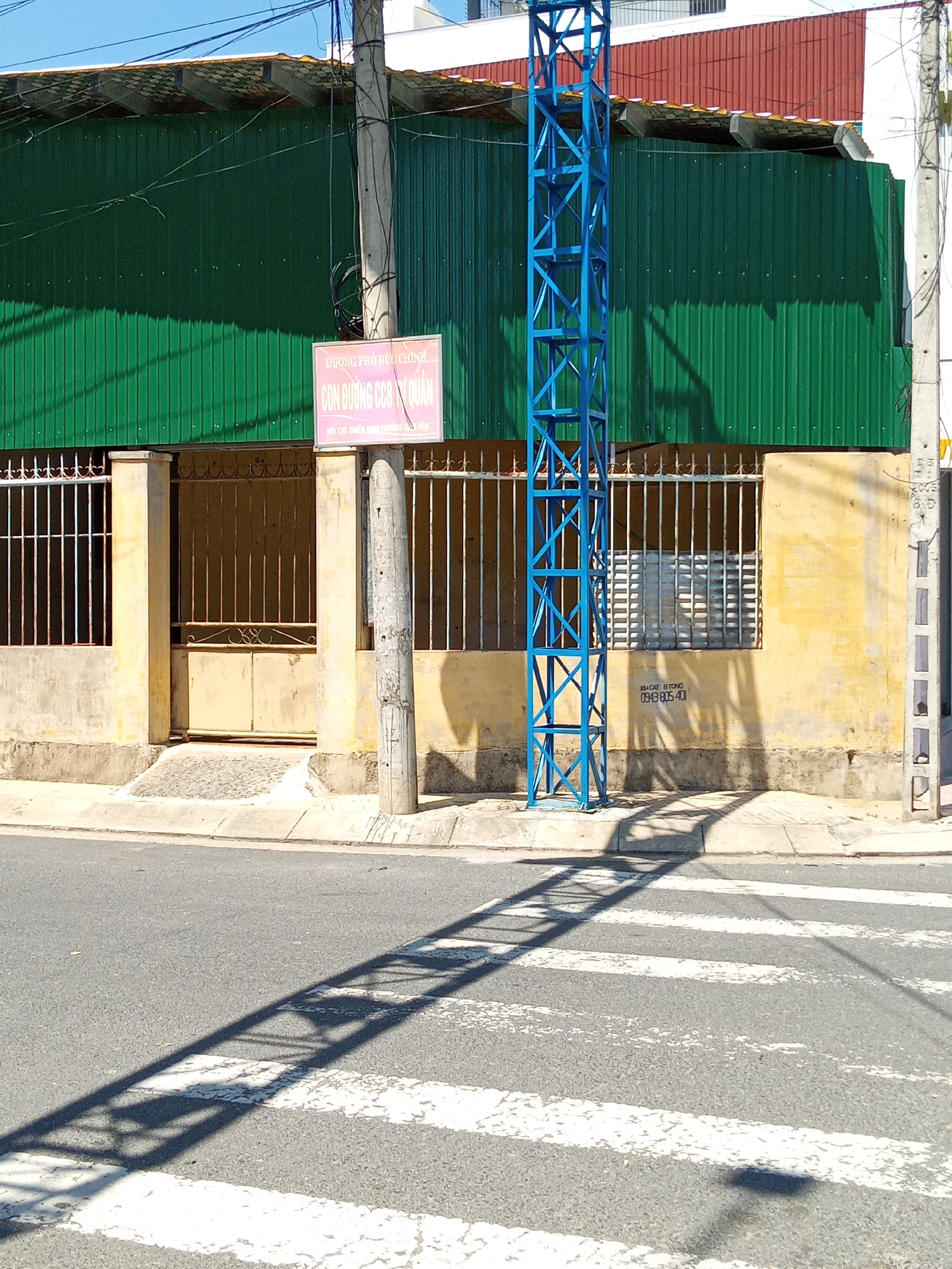 This image depicts a daytime street scene in Nha Trang, Vietnam. A two-story building with a weathered pale yellow facade and a vibrant green corrugated metal roof dominates the background. The building features a double-leaf, pale yellow metal gate, flanked by concrete pillars and barred windows, accessible via a small ramp from the concrete sidewalk. A pink rectangular sign mounted on the facade displays Vietnamese text: "DƯƠNG PHỐ ĐỨC CHÍNH", "CÓN ĐƯỜNG CCB Ý QUAN", and "HỘI CỰU CHIẾN BINH PHƯỜNG HÒA" (Veterans Association Hoa Ward). To the right of the building's entrance, a bright blue open-lattice metal structure stands on the sidewalk, casting a prominent linear shadow across the asphalt road. The road features a partially visible pedestrian crosswalk with multiple white stripes. Two concrete utility poles, heavily laden with black wires, are present: one centrally positioned in front of the building and another at the far right edge of the frame. Stenciled on the yellow wall to the right of the blue structure are the words "KH CẮT BÊ TÔNG" and a phone number "0949.805.401". A portion of a building with a reddish-brown corrugated roof is visible above the green roof. The sky is clear blue, and strong sunlight creates distinct shadows. No individuals are present in the scene.