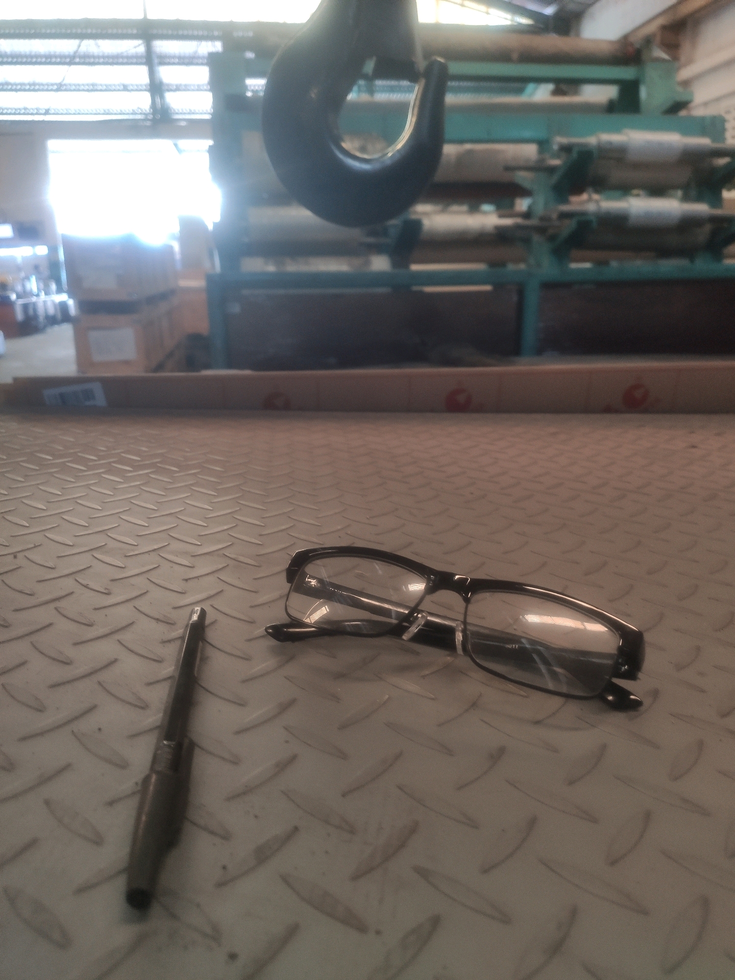 In an indoor industrial setting, a pair of black-rimmed glasses and a dark-colored pen rest on a diamond plate metal surface. The glasses are positioned to the right of the pen, with their frames and lenses visible. The pen, lying vertically on the surface, appears to be a writing instrument or a tool. In the background, industrial machinery, including a large hook and what appears to be a conveyor system or shelving unit with spools, are visible. Sunlight streams in from large windows or openings in the ceiling, illuminating the space. The overall environment suggests a factory, workshop, or storage area.