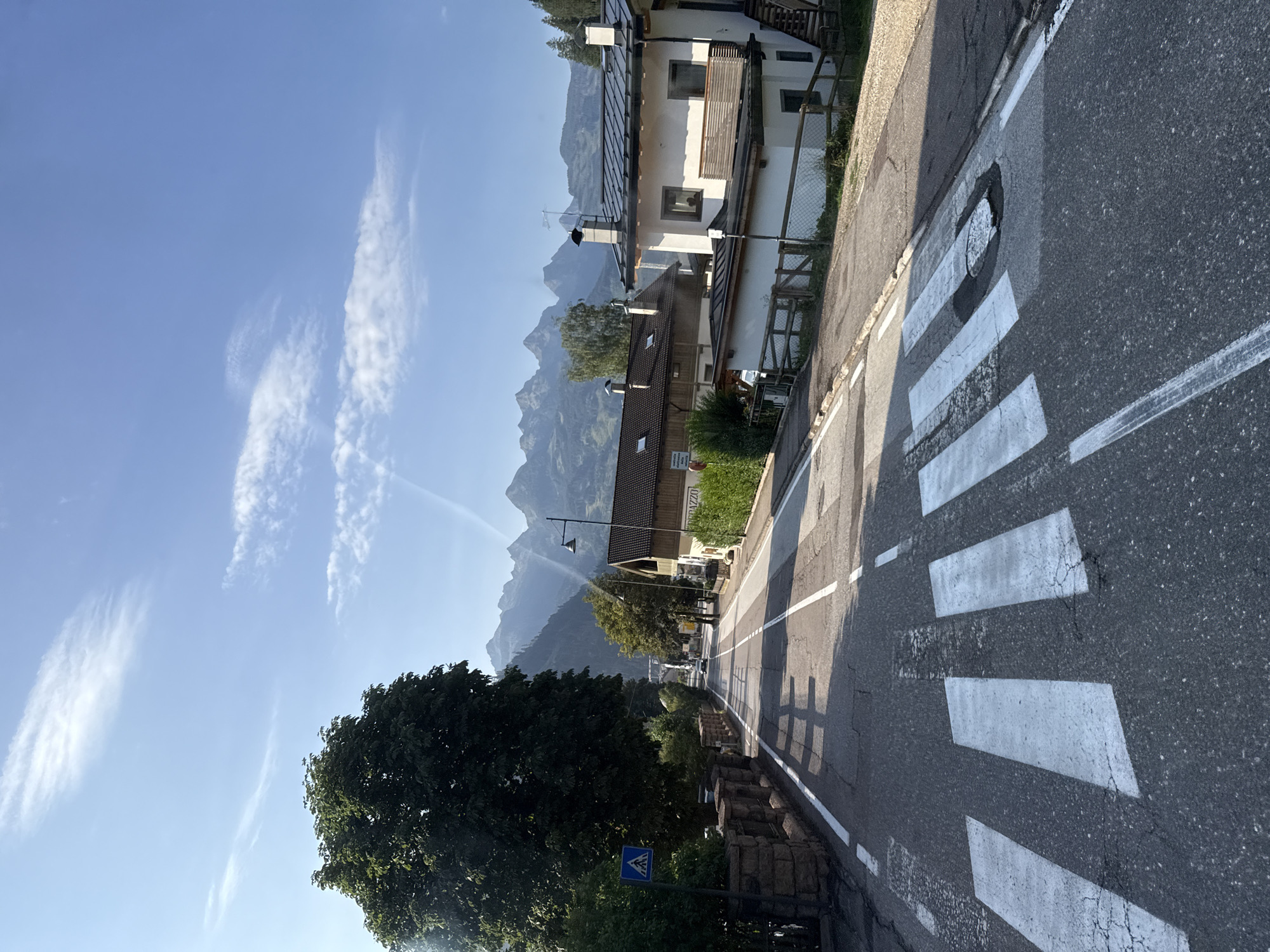 The image depicts a street scene in Predazzo, Italy, on a clear day. The foreground features a pedestrian crossing with prominent white stripes on asphalt. To the left of the road, a large, dark green tree casts a shadow on the asphalt. A stone wall borders the left side of the road, with a blue pedestrian crossing sign visible on a metal post. Further down the road, houses with traditional architecture and pitched roofs are visible, some with visible chimneys. In the background, a mountain range with jagged peaks stretches across the horizon under a bright blue sky with wispy white clouds. The road appears to be a two-lane street with white lane markings. Sunlight illuminates the scene, creating distinct shadows from the trees and buildings.