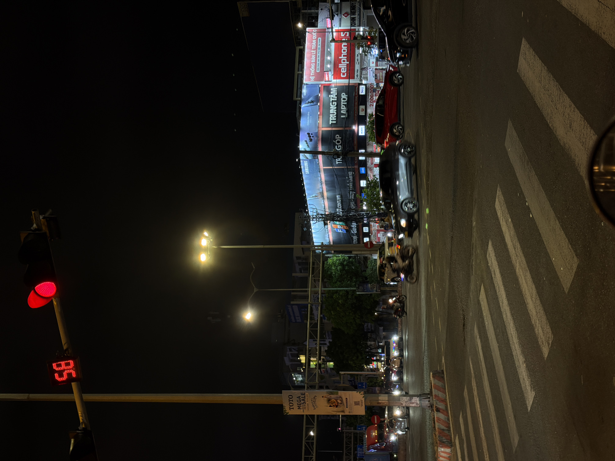 This is a nighttime street scene in Da Nang, Vietnam, likely at an intersection with traffic lights and a pedestrian crossing. A traffic light shows a red signal, with a digital timer displaying "58". Several cars and motorcycles are visible, with headlights on, suggesting traffic is moving or stopped at the intersection. Buildings with illuminated signs are in the background, including advertisements for electronics stores like "SAMSUNG", "TRUNG TÂM LAPTOP", and "cellphones". A large banner on a pole reads "TOTO MEGA SALE". The street is paved and the pedestrian crossing stripes are clearly visible. The overall atmosphere is typical of a busy urban environment at night.
