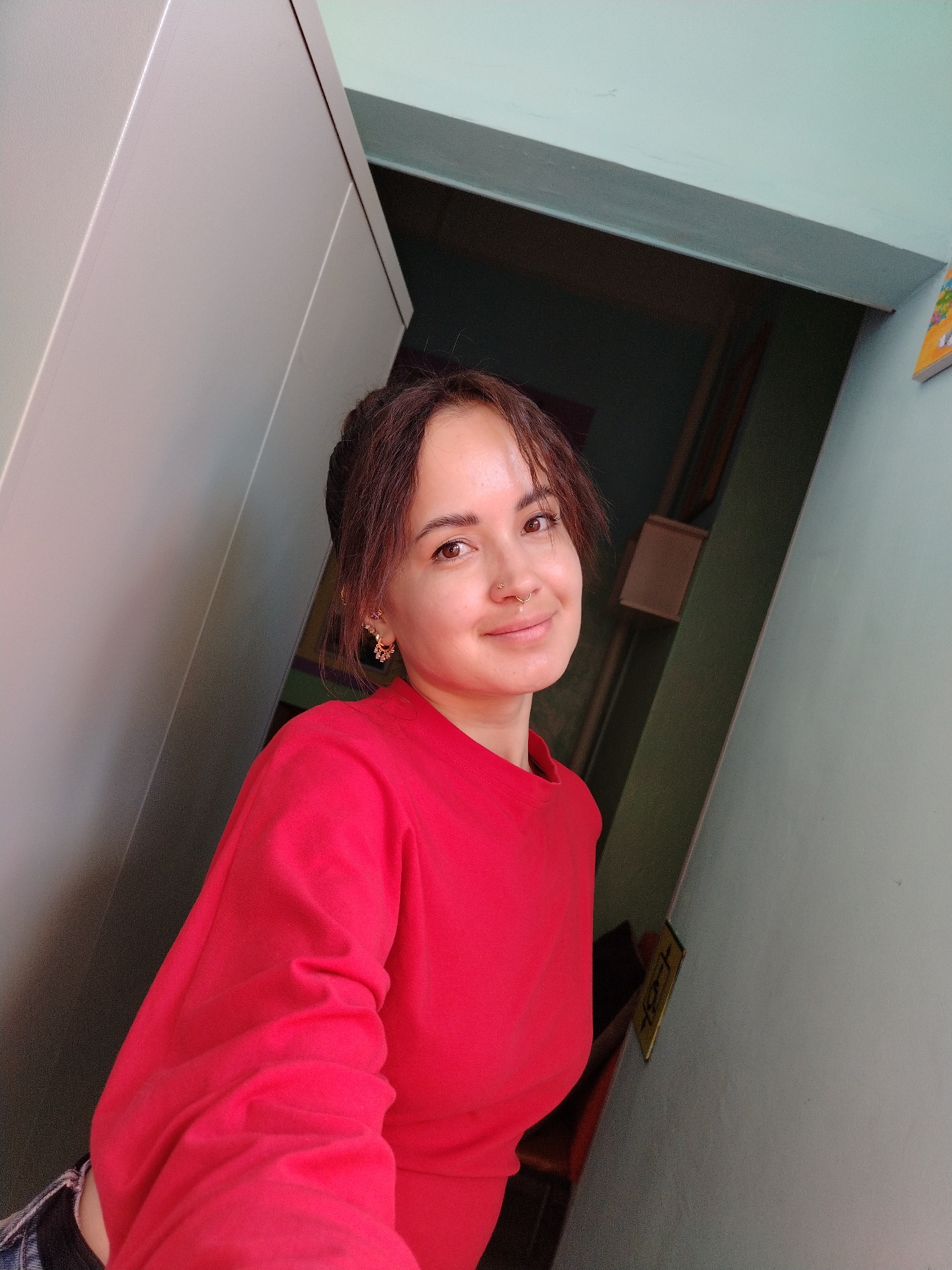 A young woman with brown hair and a nose ring is pictured in a selfie. She is wearing a red sweatshirt and blue jeans. She has on large gold hoop earrings and her hair is pulled back in a bun. The background appears to be a hallway with light blue walls and a large grey cabinet. There is a small yellow sign with black writing on the wall behind her. The image was taken in the city of Chaikovsky, Russia.