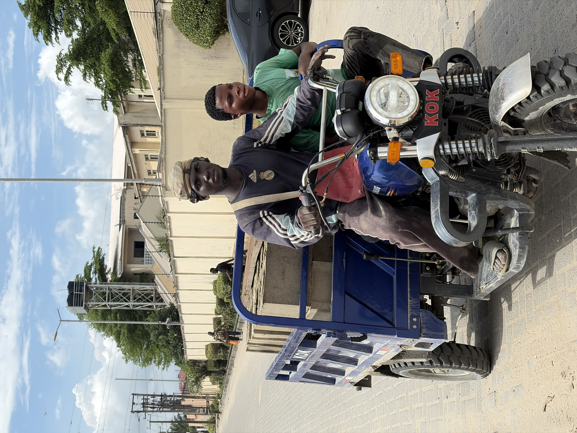 Two young men are seated on a blue three-wheeled motorcycle with a flatbed cargo area. The motorcycle has the letters "KOK" prominently displayed. The location appears to be Igboefon, Nigeria, based on context clues and visual features typical of the region. The environment includes buildings, vegetation, utility poles, and an overcast sky. One man is wearing a dark, striped shirt and a light-colored cap. The other is wearing a green shirt with striped sleeves. They are positioned in an urban area, possibly a street, with other people visible in the background.