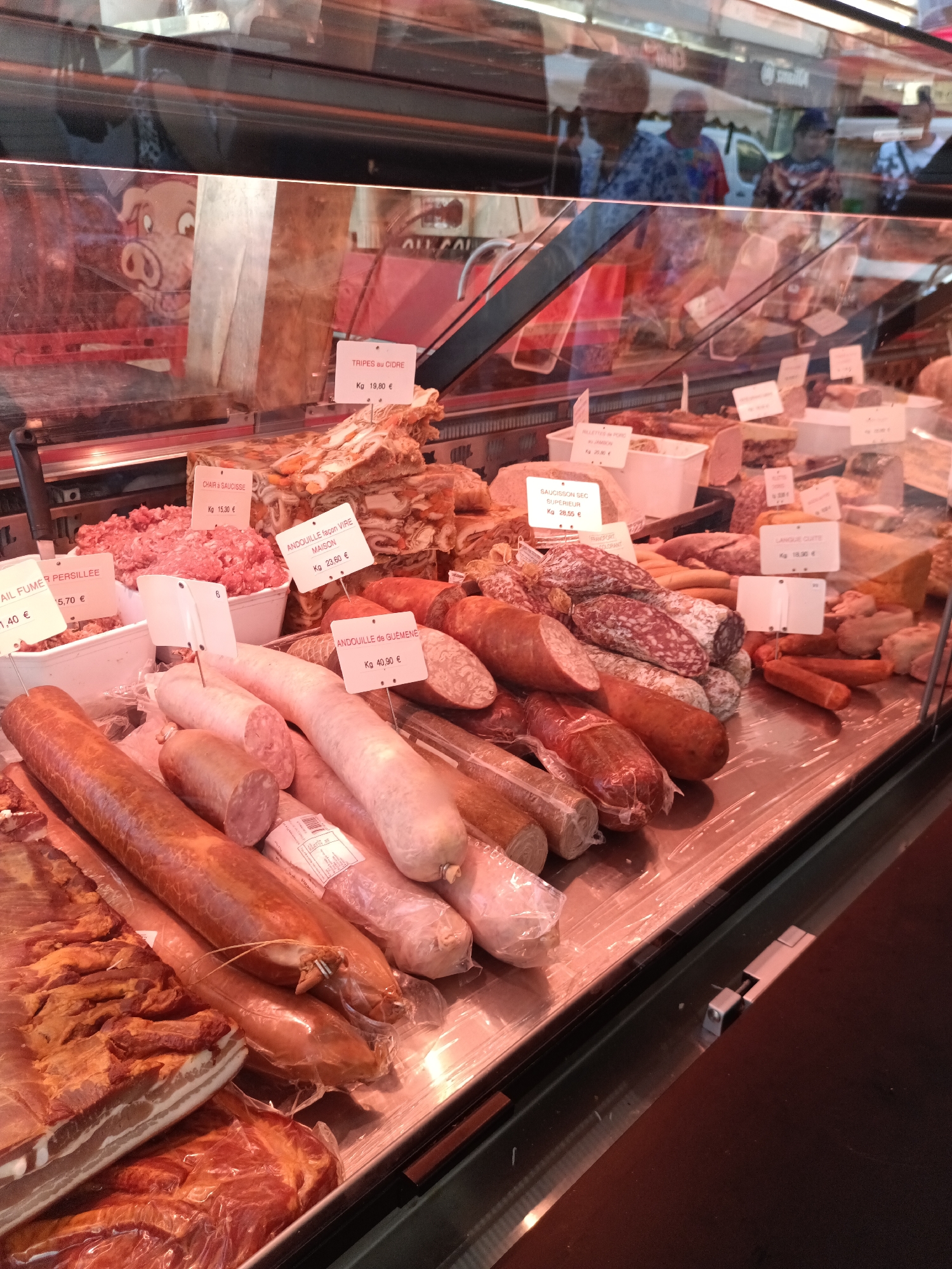 The image shows a close-up of a butcher's display case filled with various meats and sausages. The display is well-lit, showcasing a wide selection of cured meats, ground meat, and what appear to be tripe and chitterlings. Several small white price tags are visible, listing items and their prices per kilogram. Among the visible labels are "TRIPES AU CIDRE," "CHAIR A SAUCISSE," "ANDOUILLE FAÇON VIRE MAISON," "ANDOUILLE DE GUÉMÉNÉ," "SAUCISSON SEC SUPÉRIEUR," and "LANGUE CURTE." In the background, the reflection in the glass shows a market scene with people browsing, suggesting a public market setting. A cartoon pig image is also visible on a red background behind the meat. The overall impression is of a bustling food market, likely in France given the French labeling. Based on other context, this image was taken in Le Neubourg, France.