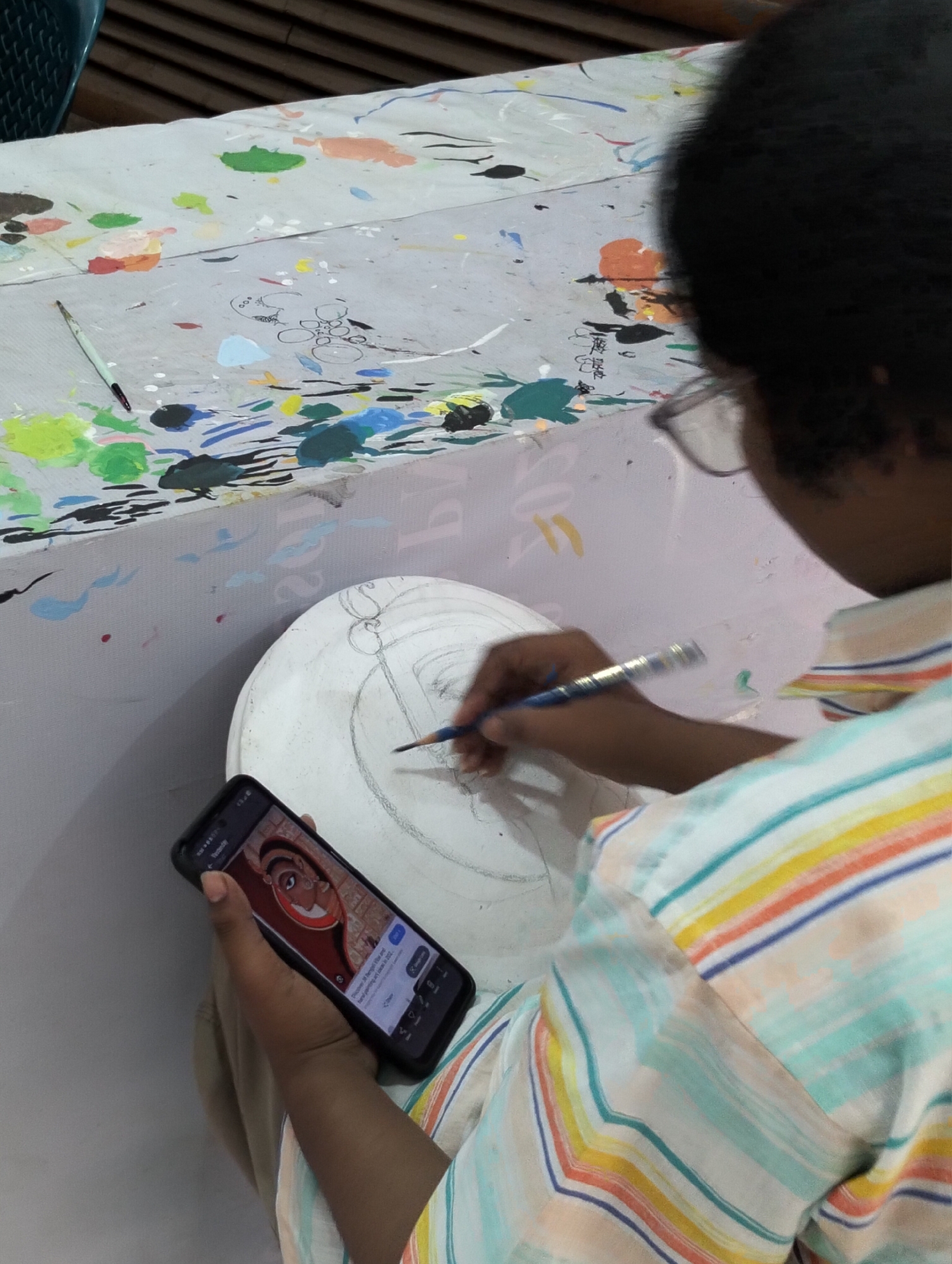 A person is seated and drawing on a round, white surface. They are holding a smartphone in their left hand displaying an image of a painting. In their right hand, they hold a pencil, drawing on the white surface. The person wears glasses and a striped shirt. A table with paint splatters is partially visible in the background, along with a paintbrush. The location is in the city of Unknown, Unknown.