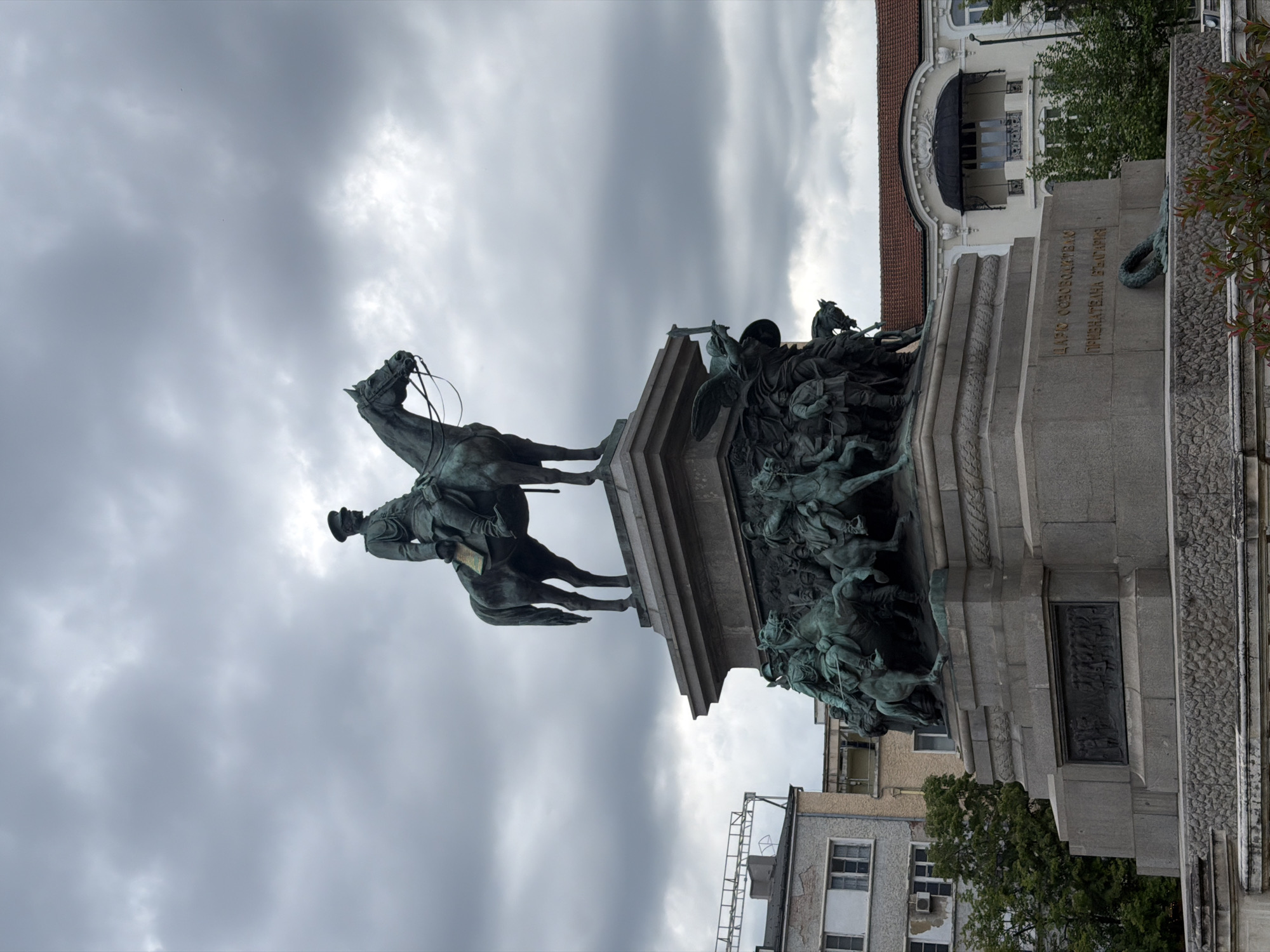 The image shows the equestrian statue of Tsar Alexander II, part of the Liberation Monument in Sofia, Bulgaria. The monument is located in the city center, surrounded by buildings and with a cloudy sky overhead. The statue depicts Alexander II on horseback, with other figures sculpted at the base of the monument. The monument's base is made of stone and contains inscriptions and bronze reliefs.