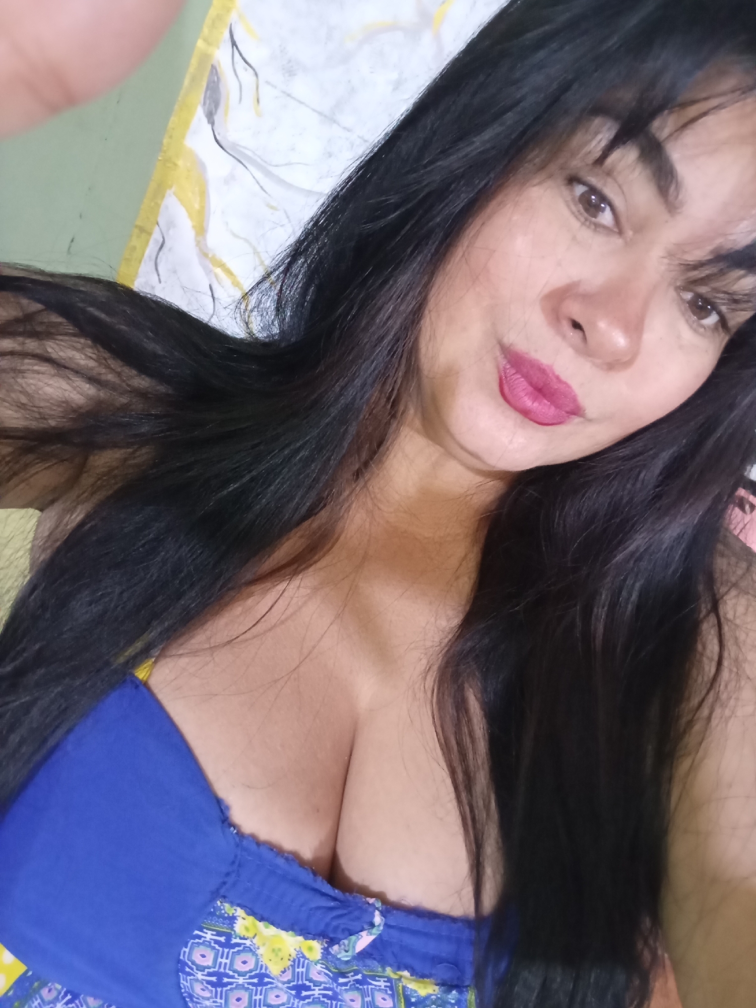 A close-up, eye-level selfie depicts a woman with long, dark, straight hair flowing around her shoulders. She looks directly at the camera with a confident, slightly smiling expression, her head tilted slightly. She has brown eyes and is wearing pink lipstick and what appears to be other facial makeup. Her hair has some bangs or fringe falling over her forehead.
She is wearing a blue top, which appears to be off-the-shoulder or strapless, with an elasticated or ruched upper edge. The lower visible part of her attire features a patterned fabric with blue, white, yellow, and pink geometric or floral designs. Her décolletage is prominent.
The background is softly out of focus. On the left, a light green or yellow-green surface is visible, while behind her head and to the right, there's a light-colored abstract pattern, possibly on a bedsheet or curtain, containing light grey and yellow elements. The lighting is soft and even, suggesting an indoor setting during the day or under artificial light. No visible text is present.
