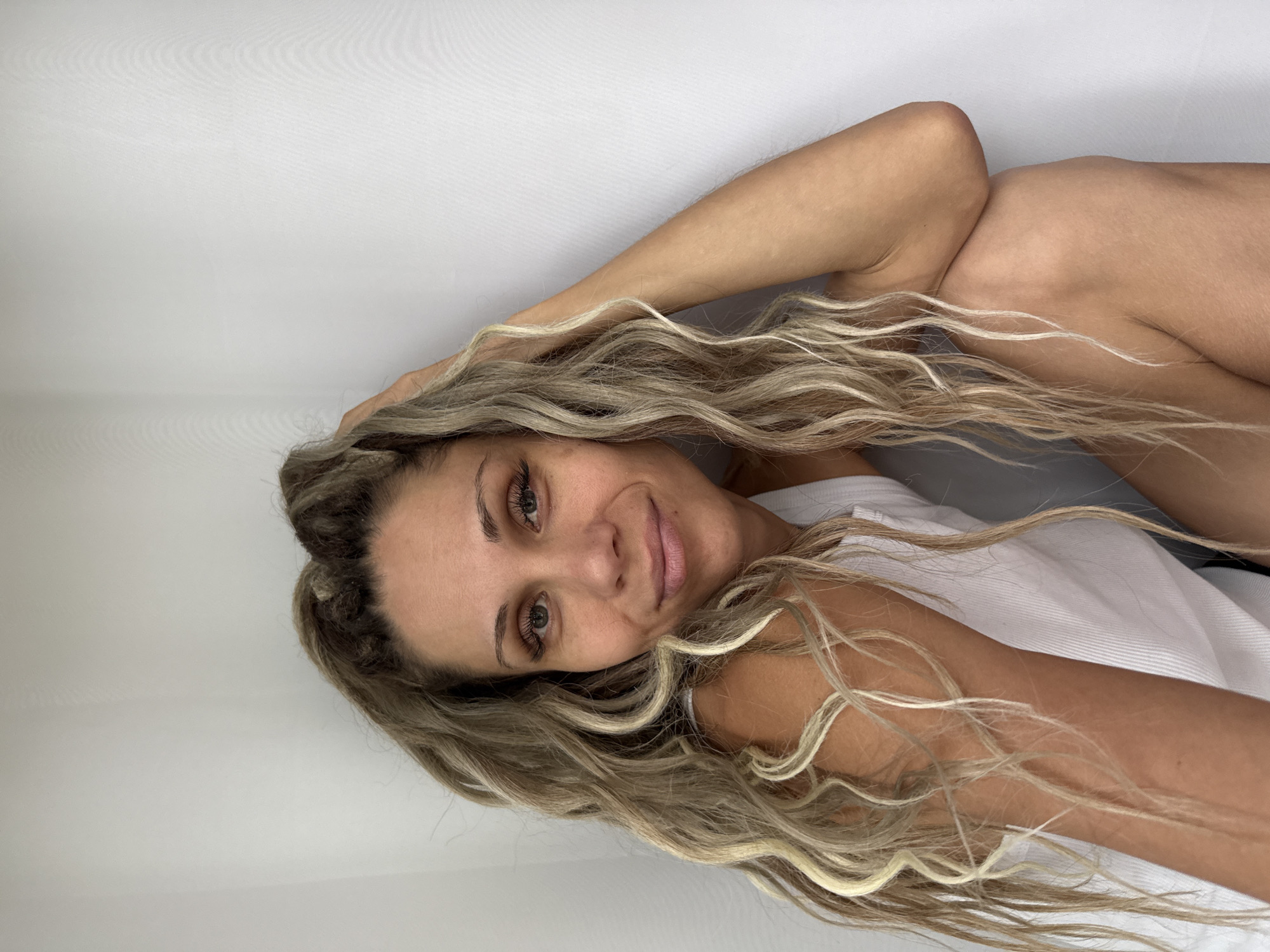 A woman with long, blonde, wavy hair is sitting with her right arm resting on her head. She is wearing a white tank top and is looking at the camera. The background is a plain white curtain. The image is a medium shot, with the woman filling most of the frame. The lighting is soft and natural, with no harsh shadows. The woman's expression is relaxed and content. There is no indication that this image was taken in Rusanovo, Russia.