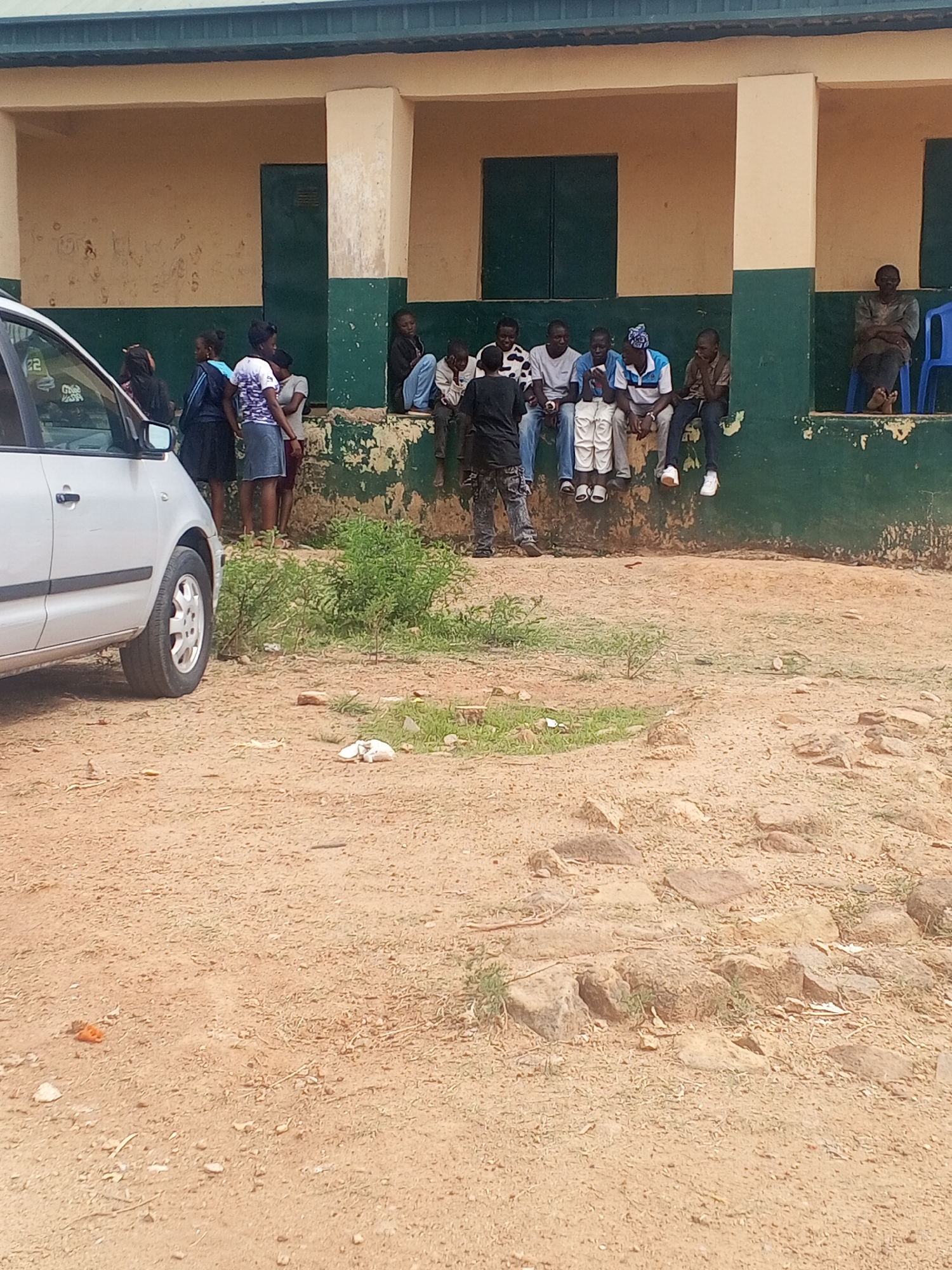 The image shows an outdoor scene in Jos, Nigeria. A silver car is parked on the left side of the frame, partially obscuring the view. In the background, there is a building with a green and beige facade. A group of people is sitting on a ledge of the building, while others stand nearby. One person is seated on a blue chair. The ground is dirt with patches of vegetation and rocks.