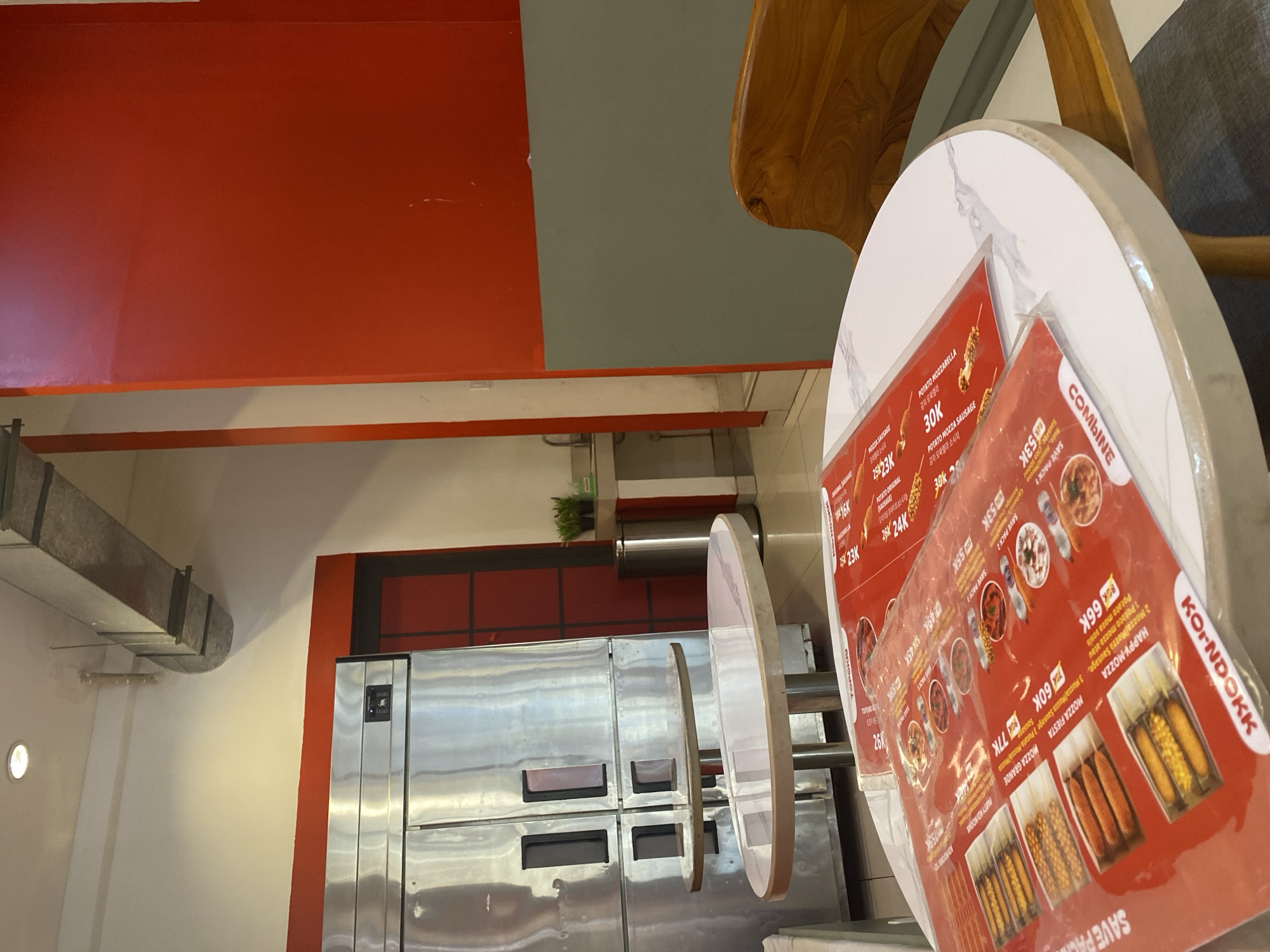 The image depicts the interior of a restaurant in East Jakarta, Indonesia, with a focus on dining tables and kitchen equipment. In the foreground, a white marble-topped round table displays two red menus with food items and prices in Indonesian currency. The menus are partially obscured by the table's edge and the viewer's perspective. One menu shows images of corn dogs and offers items like "Mozza Festa" and "Happy Mozza" for prices such as 60K and 66K.
Behind the table, two oval tables with reflective surfaces are visible, positioned in front of a large, stainless steel kitchen appliance that appears to be a multi-door refrigerator or freezer. The appliance has several dark rectangular panels on its doors. Further back, a red-painted wall provides a strong visual contrast to the metallic sheen of the kitchen equipment. A doorway with a red frame is partially visible on the left, leading to an area with a sink and faucet, suggesting a food preparation or washing station.
The environment is well-lit, with overhead lighting visible on the ceiling, and a metal ventilation duct runs horizontally across the upper part of the image. A small potted plant sits on a counter next to a silver trash can in the background. A wooden chair is partially visible on the right side of the frame, indicating seating arrangements. The overall scene suggests a modern and clean dining establishment, likely a fast-casual restaurant specializing in street food like corn dogs, given the menu contents.