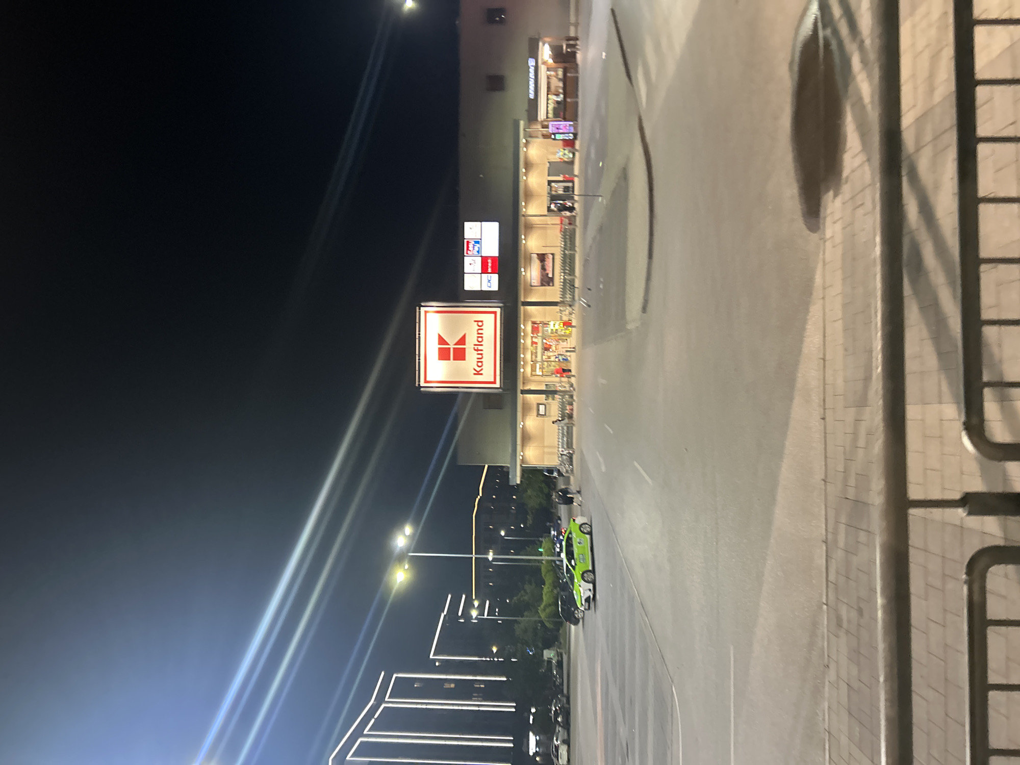 The media file captures a commercial area at night in Plovdiv, Bulgaria, characterized by artificial illumination. The central focus is a large building, likely a shopping center or supermarket, with several illuminated signs. Most prominent is a red and white sign for "Kaufland" at the bottom left, clearly displaying the brand name and logo. Above it, other smaller, brightly lit signs are visible; one appears to read "Secomania" and another includes the text "DC Redi."
In front of the building, there is a paved road or parking lot where a bright green compact car is distinctly visible, along with other parked vehicles further in the distance. Shopping carts are lined up near an entrance to the building. The scene is illuminated by numerous streetlights, which create prominent light streaks across the dark sky, indicating a night-time capture, possibly with a long exposure or motion blur.
To the left of the main building, modern structures feature distinctive horizontal strip lighting, contributing to the urban night ambiance. No people are clearly visible, and no specific event or activity is taking place beyond the general function of a commercial area. The overall impression is that of a bustling, well-lit commercial district after dark.