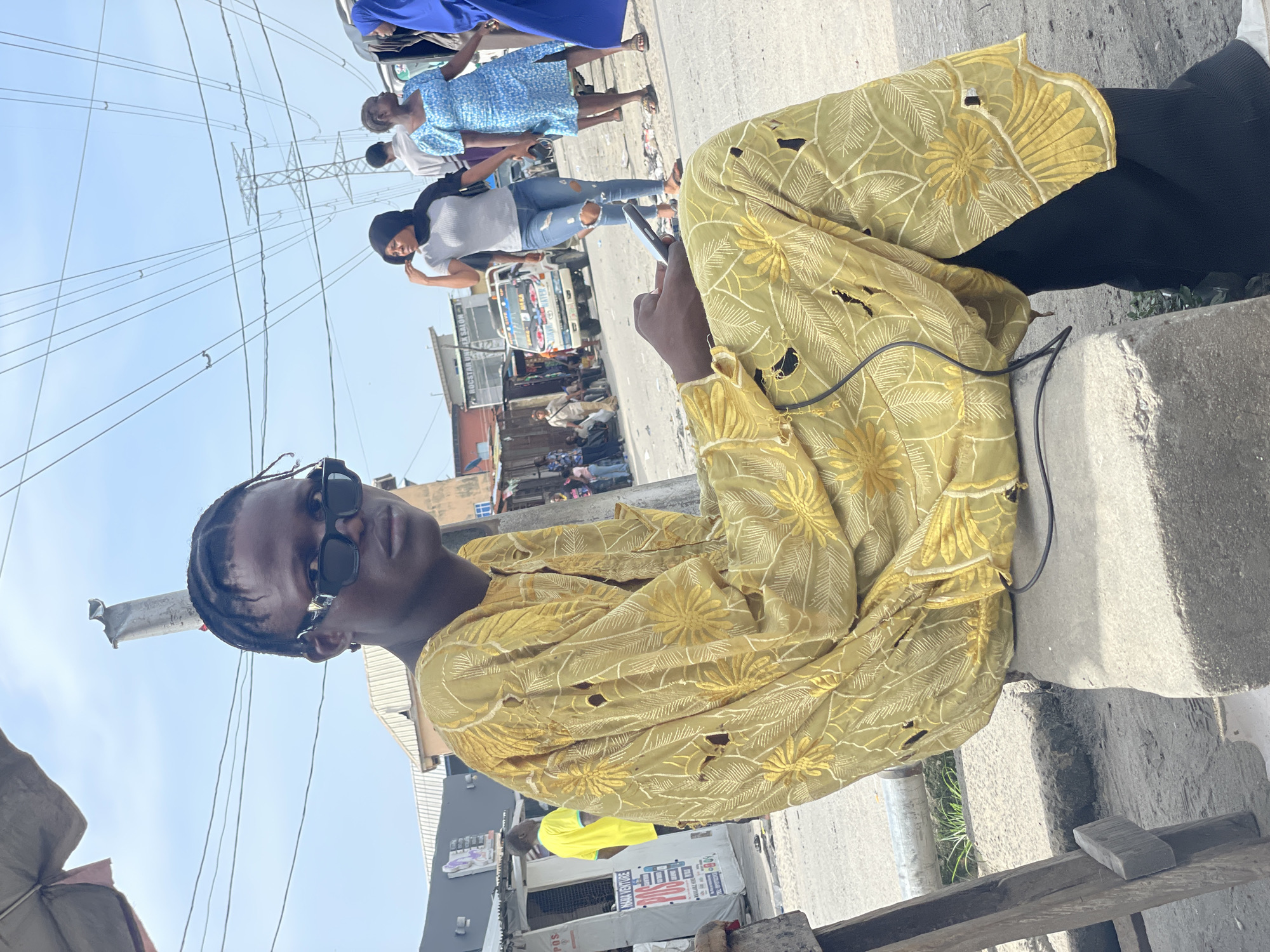 A medium, tilted, outdoor shot captures a person wearing sunglasses and a yellow, floral-patterned, long-sleeved top with holes, sitting on a concrete structure on the side of a street. The individual is looking at the camera while holding a phone in their hand. In the background, there are people walking along the street, and commercial buildings can be seen, indicating an urban setting. The presence of power lines overhead suggests the image was taken in a populated area. Based on the prompt, the location is Ado, Nigeria.