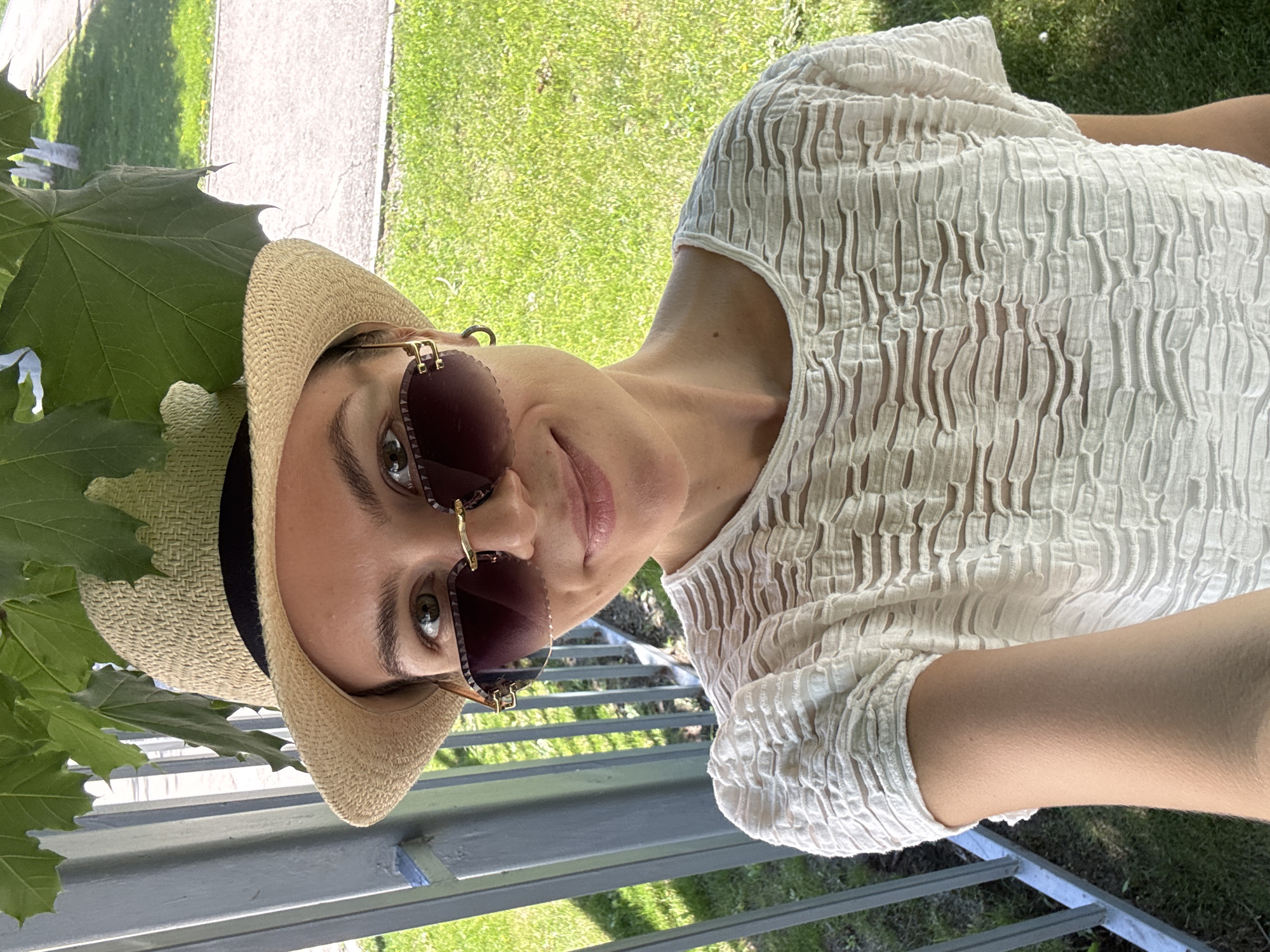 A woman wearing a hat and sunglasses is standing outside near a railing. She is wearing a white textured shirt. Green grass and a sidewalk can be seen in the background. The location is Unknown, Unknown.
