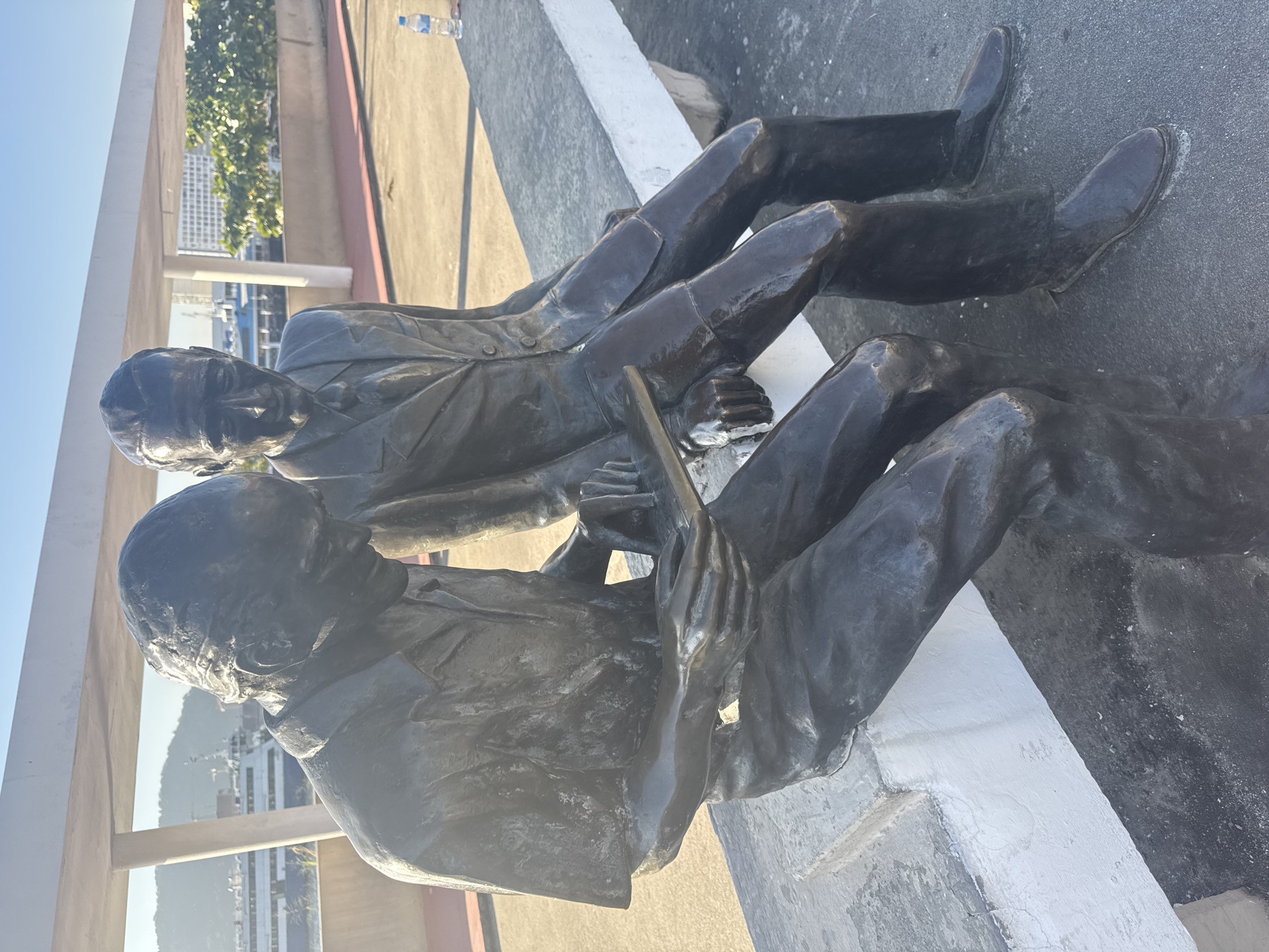 The image shows a bronze sculpture of two men sitting on a concrete bench. The man on the left is looking down at a tablet held in his hands, while the man on the right looks forward. They are both dressed in suits. The sculpture is located in Niterói, Brazil, near the MAC (Museu de Arte Contemporânea), which can be seen in the background. The setting appears to be an outdoor promenade or park with a view of the bay and city. The sky is clear and blue, indicating a sunny day. A water bottle is visible on the bench to the right of the sculpture.