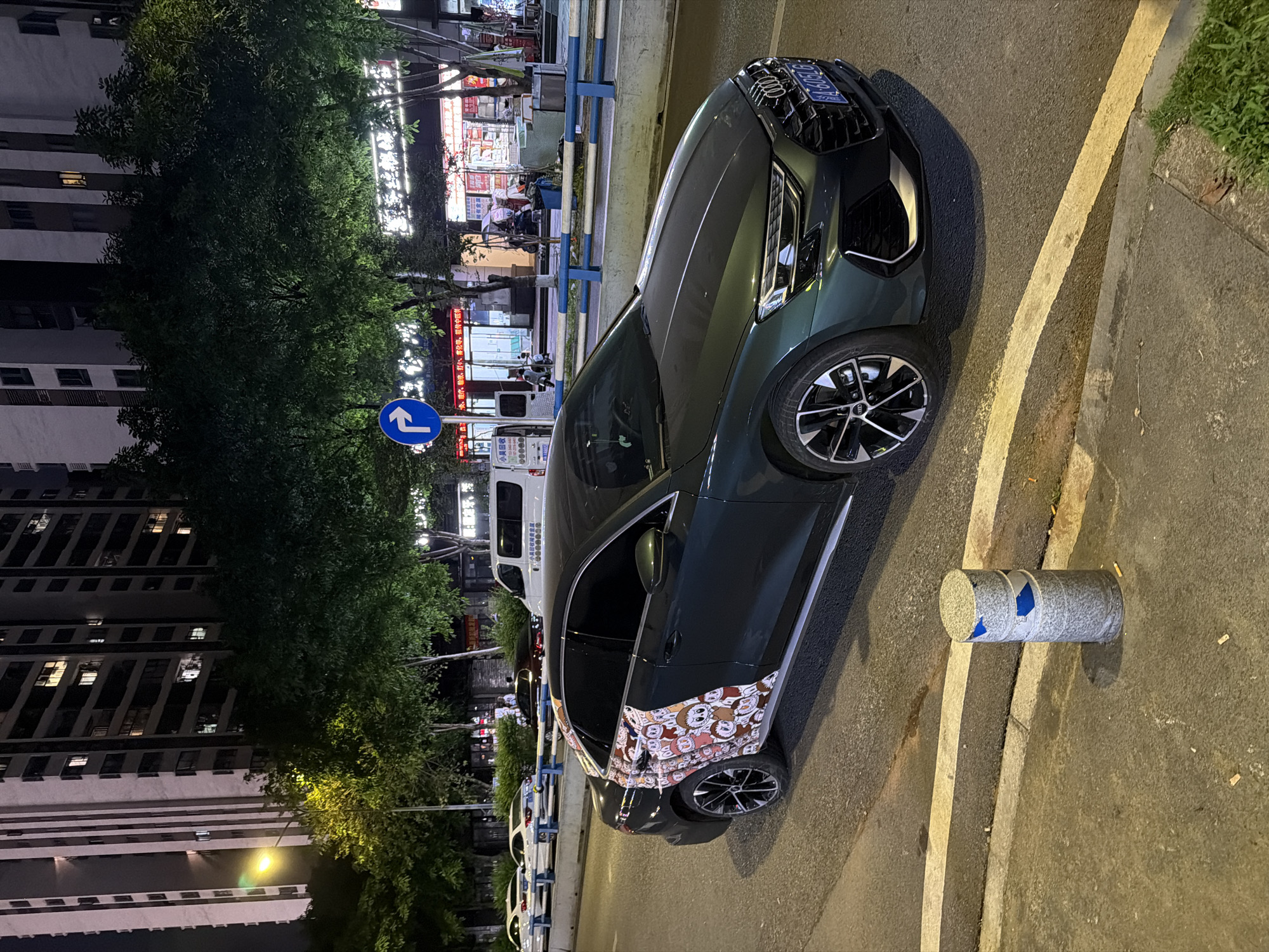 The image captures a nighttime scene in the Chongqing High-tech Zone, China. A dark green Audi sedan is parked on the right side of the street, near a cylindrical concrete barrier with remnants of blue tape. The car's rear side window is decorated with cartoon character stickers. In the background, a street lined with trees and buildings is visible. Storefronts with bright signage illuminate the street, and a blue directional sign indicates a left turn. A white van and several other vehicles are parked further down the street. A tall apartment building with numerous lit windows is seen on the left side of the image.