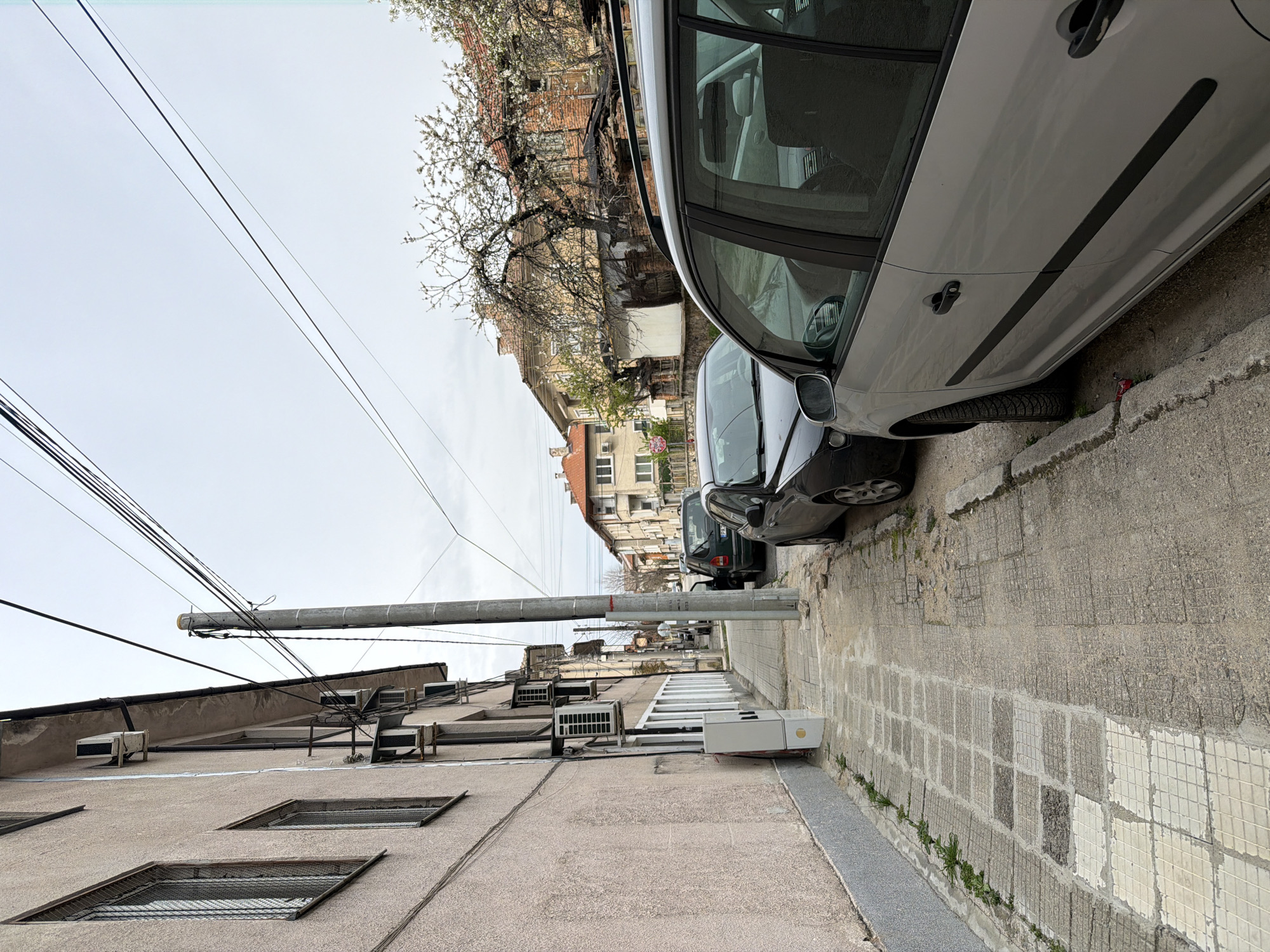 A narrow streetscape in Panagyurishte, Bulgaria, is captured with a low-angle perspective. The view is dominated by buildings on either side and parked cars along the right curb. The building on the left exhibits a faded beige exterior, interspersed with small, barred window openings and multiple air conditioning units mounted near the top. Above, a dense network of overhead wires intersects with a concrete utility pole.
Parked vehicles, including a silver car and a dark-colored car, occupy the right side of the street. The sidewalk is paved with square concrete slabs. In the background, buildings with terracotta roofs and blooming trees are visible against a cloudy sky.