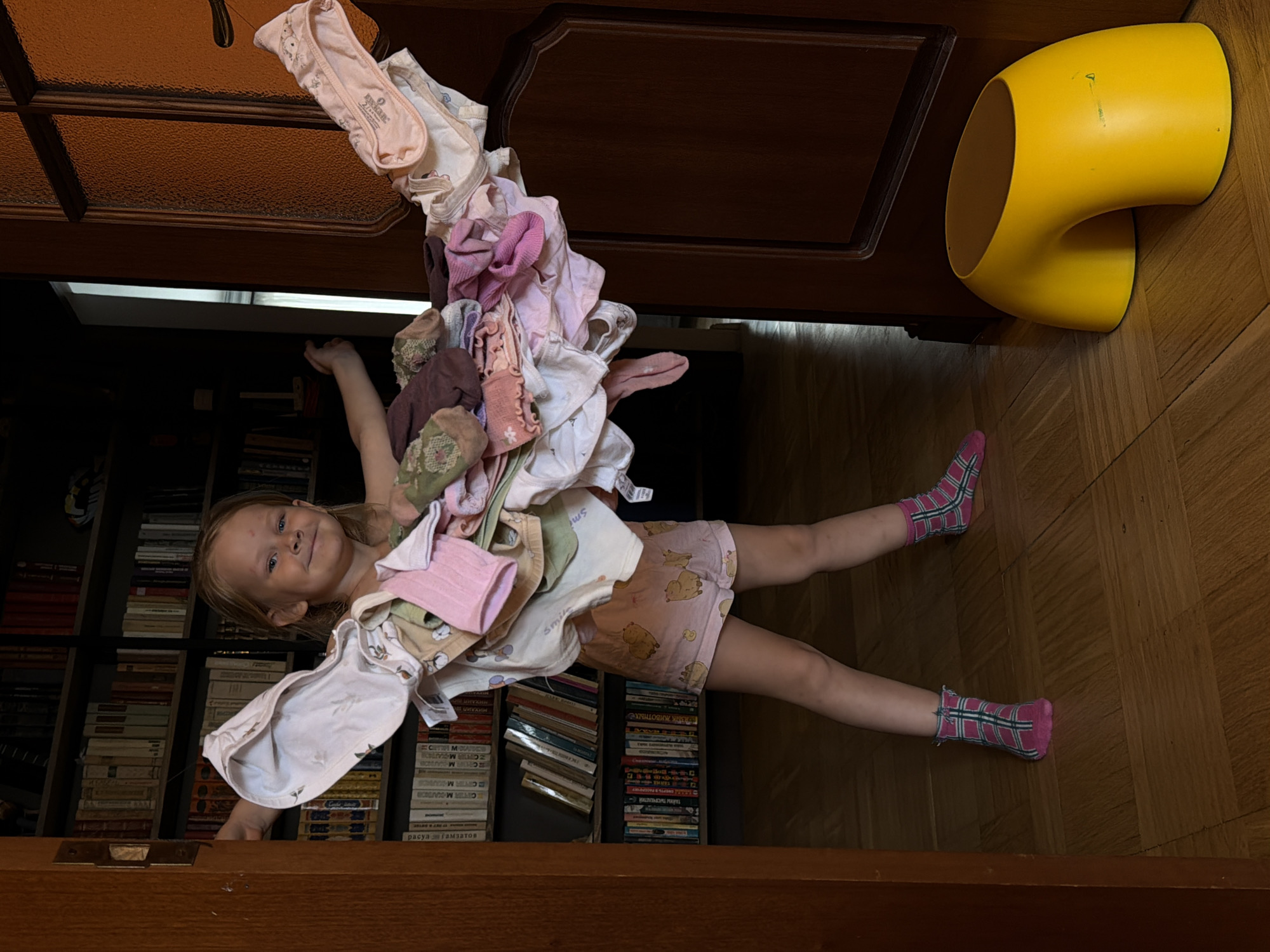 A young girl with blonde hair is playfully draped over a doorway, partially obscured by a cascade of children's clothing. She is wearing pink plaid socks and light-colored shorts with cartoon characters. Her left hand is extended, reaching towards a bookshelf filled with books. The background reveals a wooden floor and a dark wooden door frame. To the right of the girl, a bright yellow, curved object, possibly a piece of furniture or a toy, is visible. The lighting suggests it is daytime indoors. The scene conveys a sense of lighthearted mischief and a child's imaginative play. The visible text on the books appears to be in Cyrillic script.