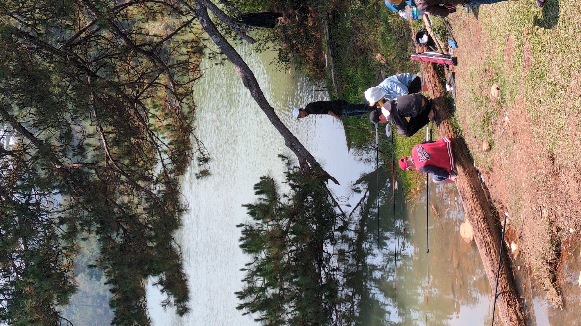 The image shows a group of people fishing in a lake in Lang Biang, Da Lat, Vietnam. Several individuals are visible, some sitting on a fallen log near the water's edge, holding fishing rods. There are trees lining the bank of the lake, and the water appears calm. Fishing equipment and other items are scattered on the ground around the people. The surrounding environment consists of grassy areas and trees, with the lake extending into the distance.