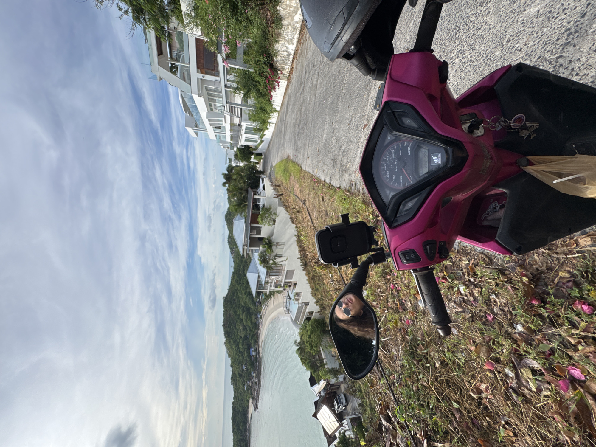 The image captures a first-person perspective from a pink scooter, parked on a paved road overlooking a tropical coastline. The scooter's handlebars, dashboard with a speedometer, and a rearview mirror are prominent in the foreground. The rearview mirror reflects a person wearing sunglasses, looking towards the camera.
Beyond the scooter, a steep, tree-covered hillside slopes down to a sandy beach and turquoise water. Several modern white buildings, seemingly villas or apartments, are nestled on the hillside, some with balconies and pools. The sky is a bright blue with scattered white, wispy clouds, suggesting a clear day.
The presence of lush greenery, the architecture of the buildings, and the tropical beach environment strongly indicate a warm-weather, possibly exotic location. The lighting suggests it is daytime, with the sun casting light and shadows. The overall mood is relaxed and inviting, hinting at a holiday or travel experience.
The visible text on the scooter's dashboard includes the "HONDA" logo and a speedometer with markings up to 160 km/h. There are also various symbols and indicators on the dashboard. A set of keys is visible hanging from the ignition. The ground around the scooter is covered in dry grass and fallen pink flowers.