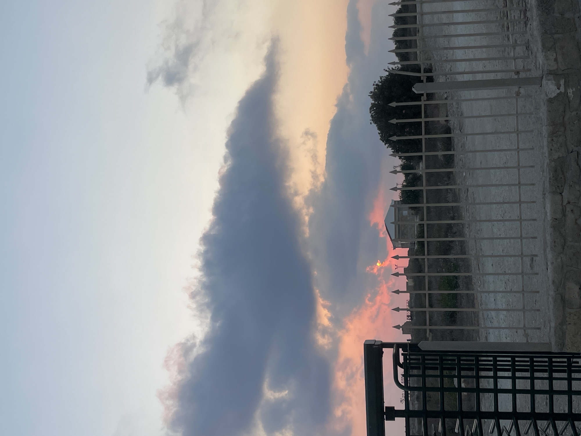 An outdoor, eye-level shot captures a dusky sky with prominent, dark grey clouds illuminated by the setting sun, which casts a vibrant pink and orange glow through gaps in the clouds. In the foreground, a black metal turnstile gate stands to the left, partially obscuring a light-colored metal fence with pointed finials that stretches across the image. Beyond the fence, a sparse, dry landscape with scattered greenery and a few small structures is visible. One building, with a light-colored facade and a dark roof, is situated on a slight rise in the middle ground. The overall impression is one of a rural or undeveloped area on the outskirts of Paphos Municipality, Cyprus, at dusk.