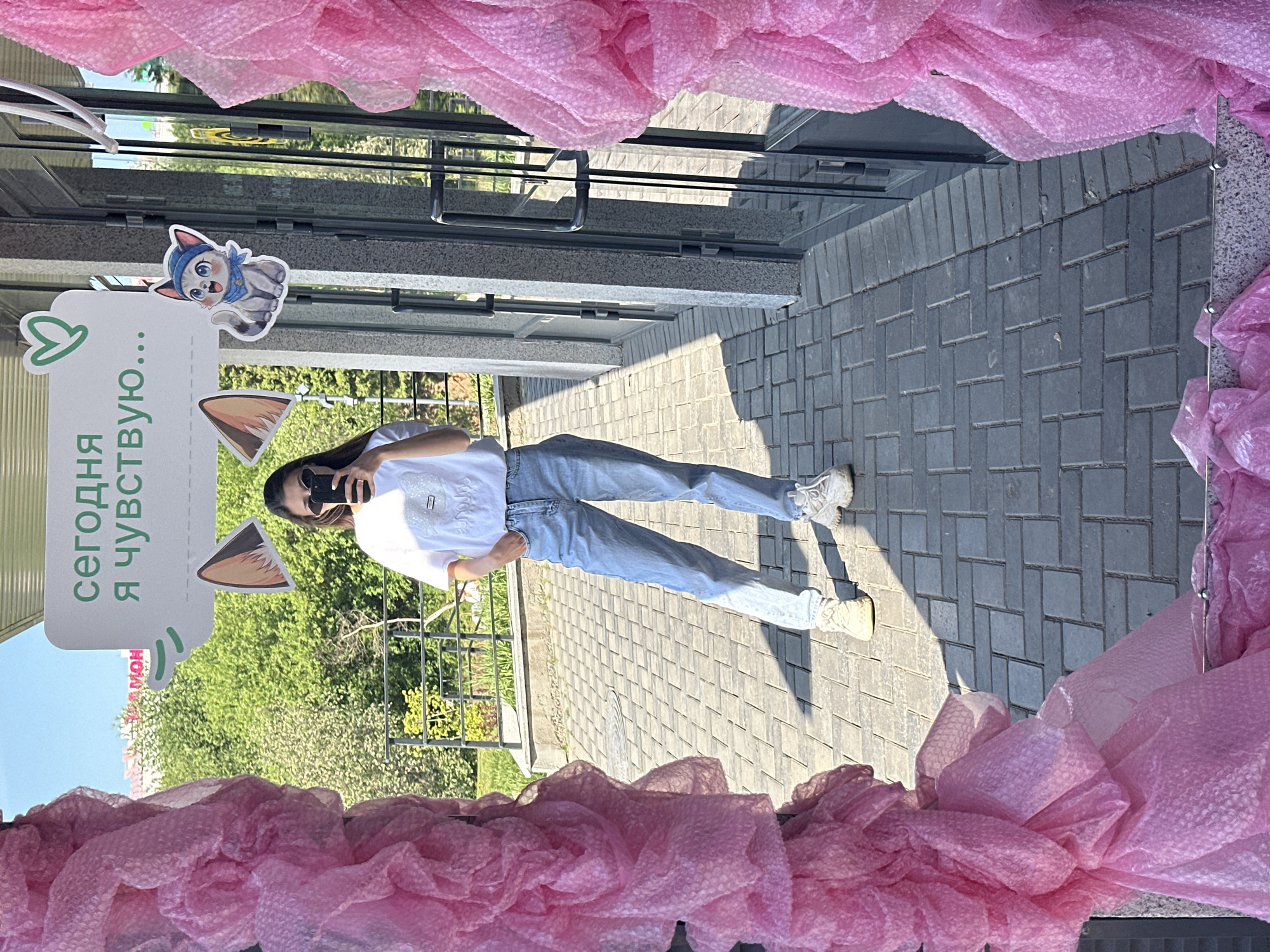 A woman in jeans and a white t-shirt is standing on a brick sidewalk, reflected in a storefront window. She is holding a phone to take a picture. In the window, there's a sign with text in Russian ("сегодня я чувствую...", meaning "today I feel..."), cat ears, and a cartoon cat image. The storefront window is decorated with pink bubble wrap. The location is Izhevsk, Russia. A building is across the street. The sidewalk leads to a building with a glass door. There is vegetation in the background.
