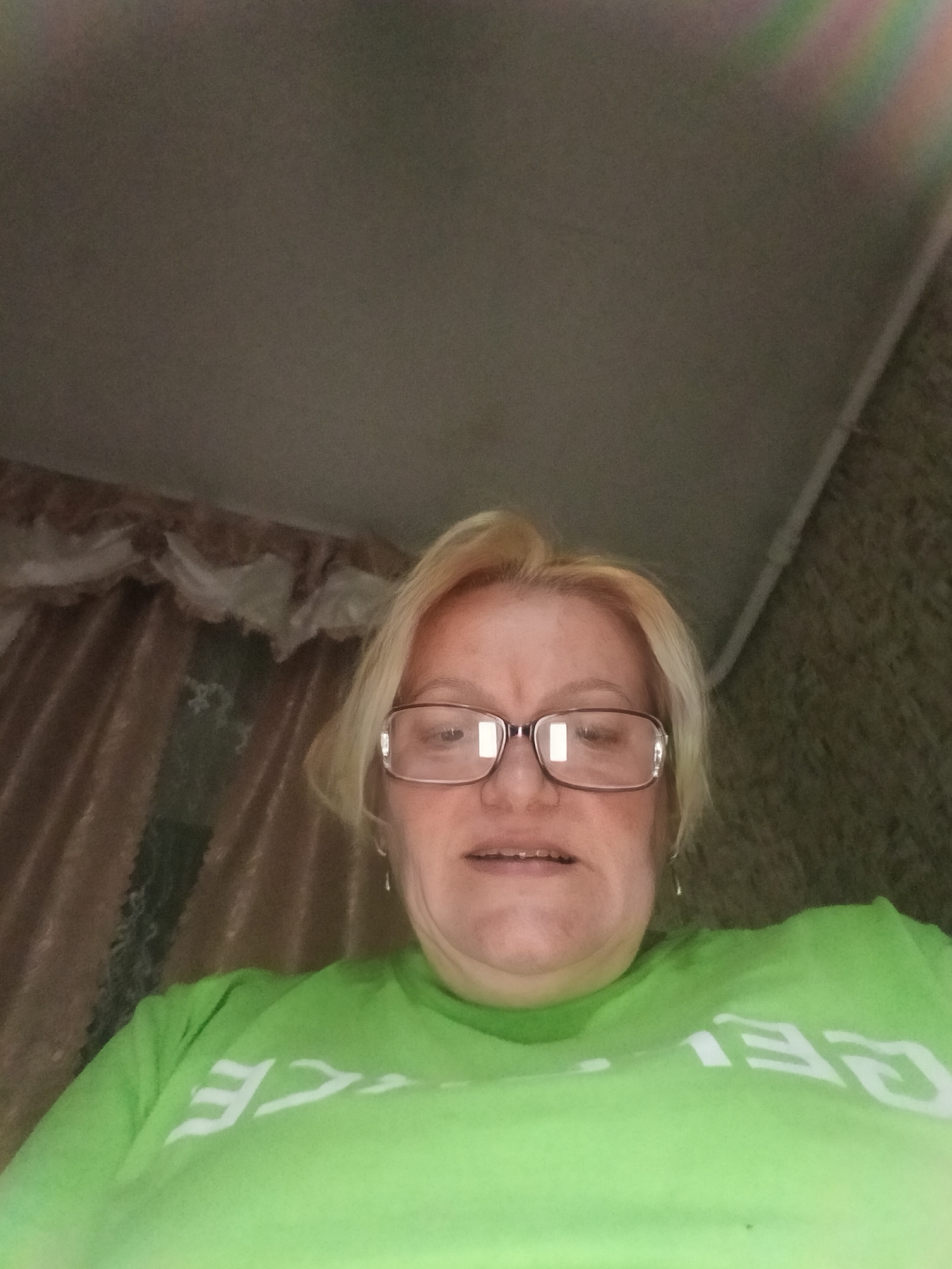 The image shows a woman wearing glasses and a green t-shirt. She has blond hair and is positioned indoors. Behind her, there is a ceiling and what appears to be a curtain or wall covering. The location is Красный Маяк, Russia.