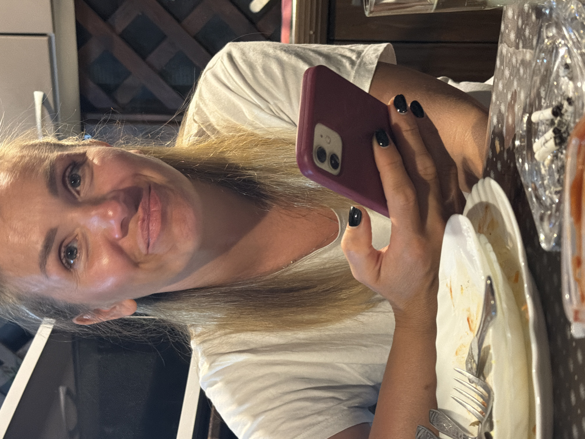 In Sochi, Russia, a woman with blonde hair pulled back sits at a table. She wears a white t-shirt and holds a burgundy smartphone in her left hand. Her nails are painted black. In front of her, a dirty white plate with leftover food and utensils is visible. An ashtray containing cigarette butts is also on the table, along with a glass of liquid. The background suggests an outdoor or semi-outdoor seating area with wooden lattice work.