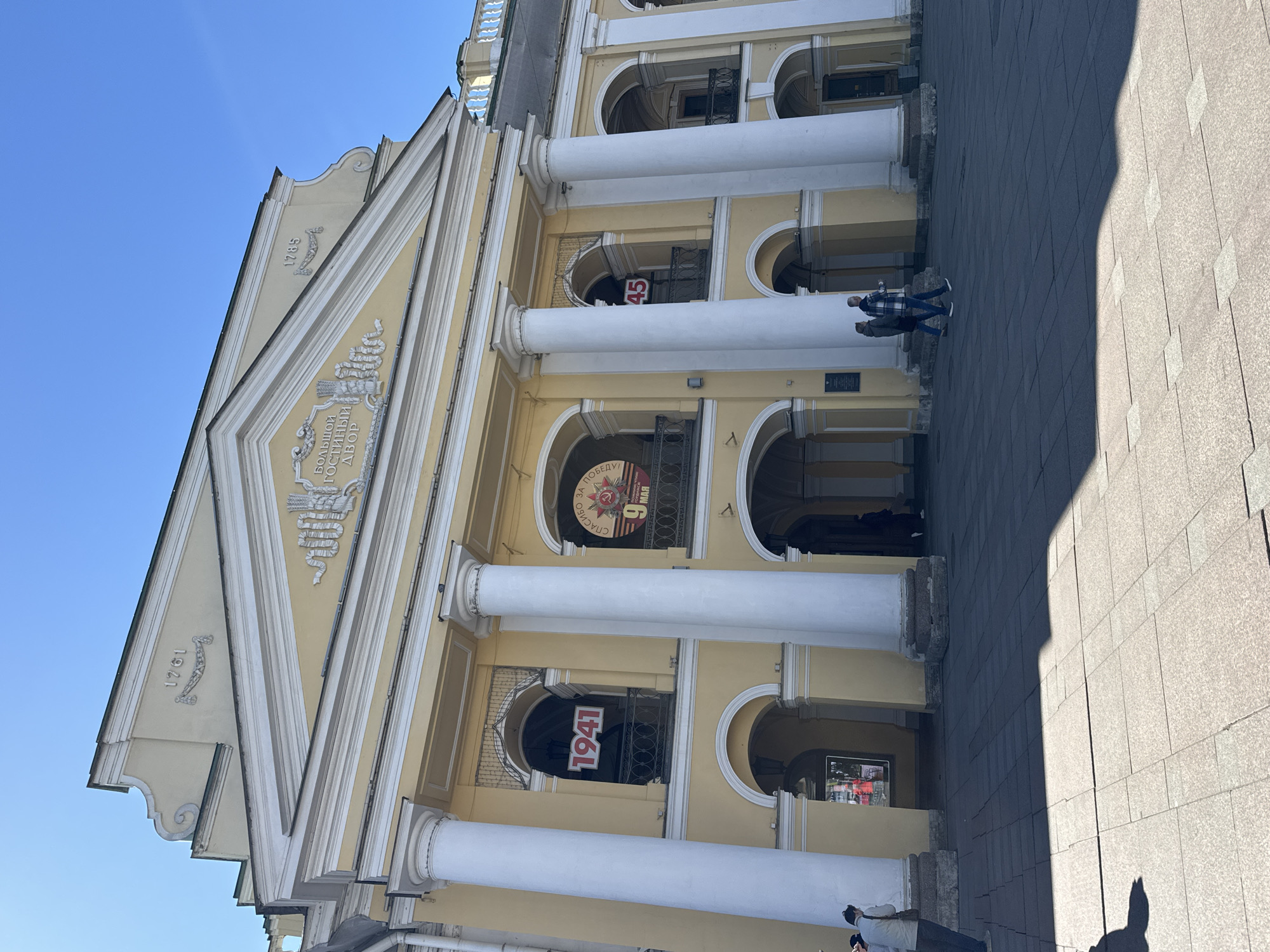 The image shows the facade of the Grand Gostiny Dvor in Saint Petersburg, Russia. The building is yellow with white columns and a triangular pediment. The date "1785" is displayed on the left side of the building, and "1961" is displayed on the left side. The phrase "Большой Гостиный Двор" is above the entrance. Posters with the dates "1941" and "45" are displayed in the windows. Two people are visible walking down the steps in front of the building. The sky is blue.