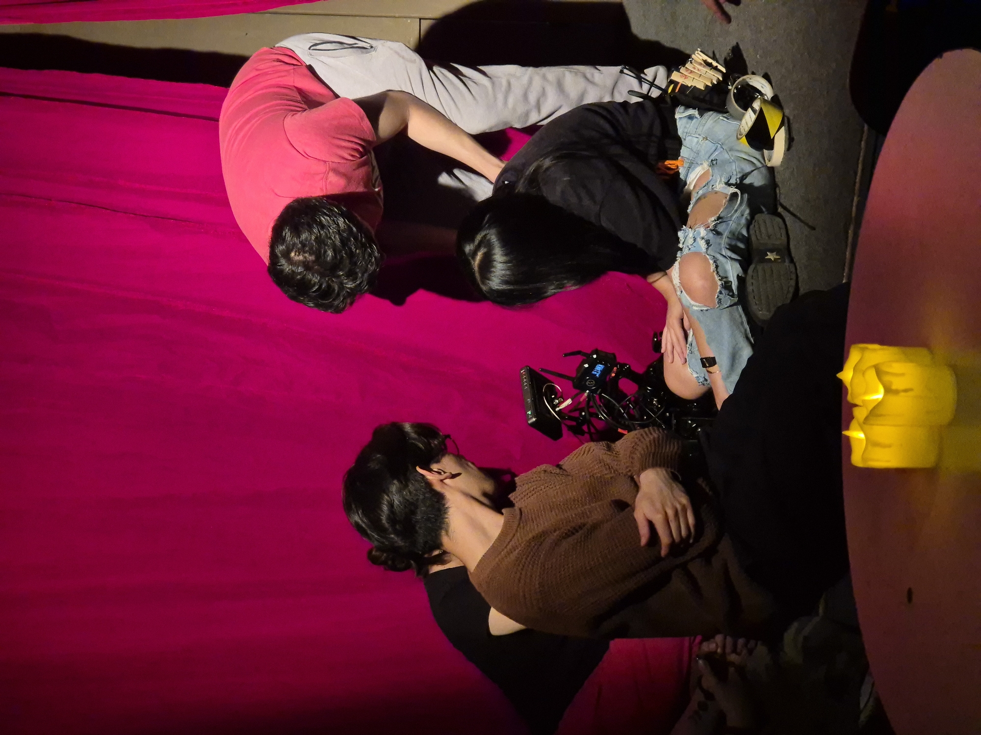 Three individuals are engaged in a production setup, likely filming or photography, against a vibrant magenta fabric backdrop. The scene is dimly lit, suggesting an indoor or evening setting.
In the foreground, a person with dark hair, wearing a brown knitted sweater and dark bottoms, is intently focused on a professional camera rig. Their hands are near the equipment, suggesting operation or adjustment. The camera has wires and a small screen displaying unreadable blue text/graphics.
To the left of the camera, a second person with long dark hair, wearing a black long-sleeved top and ripped light blue jeans, is also bent over and facing downwards, appearing to interact with or observe the equipment. Near their feet are a dark shoe with a star pattern on the sole, a roll of tape (black and yellow), and what appears to be wooden clothespins or clamps.
Further to the left, a third person with dark hair, wearing a pink/red t-shirt and grey sweatpants, is bent over the magenta fabric. Their posture suggests they are part of the crew, possibly assisting or positioning something.
On the right side of the image, a reddish-pink table is visible with decorative yellow candle-like objects that emit a soft glow. The overall atmosphere indicates a concentrated effort in a staged or studio-like environment in Puebla, Mexico.