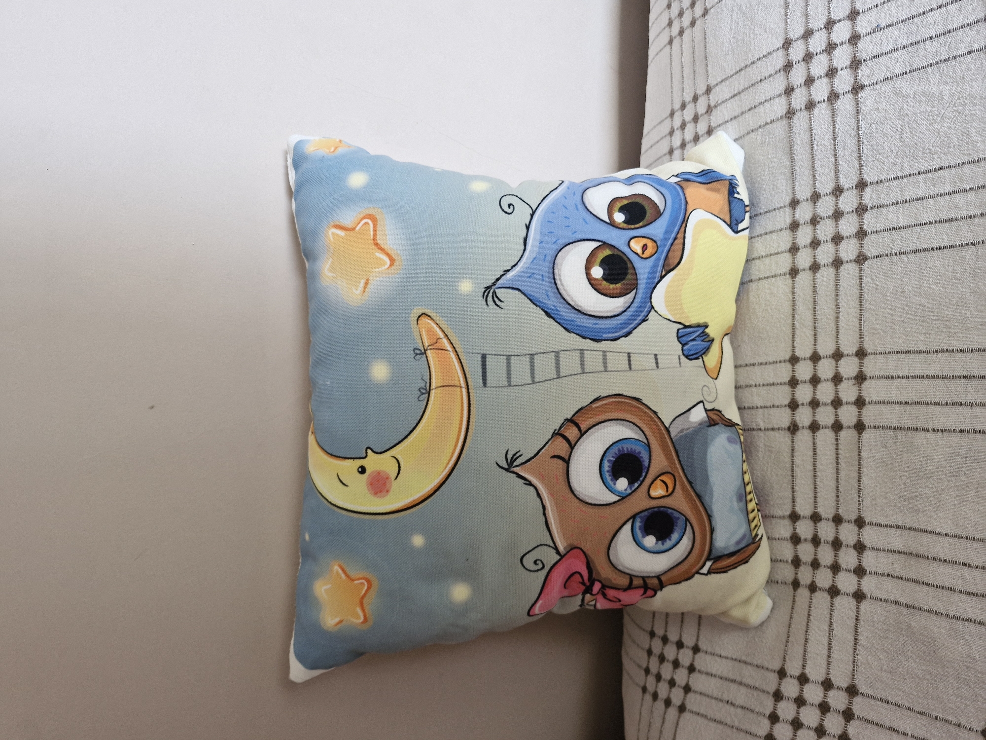 A square-shaped pillow with a cartoon illustration of two owls and a smiling crescent moon is placed on a surface covered with a beige and brown plaid fabric. The pillow's background is a gradient of light blue to darker blue, with yellow stars and glowing dots scattered across it. A crescent moon with a smiling face is positioned in the upper right portion of the pillow. Below the moon are two owls, one brown and one blue, each with large, wide eyes. The brown owl wears a pink bow on its head. A ladder-like structure is depicted between the owls, extending towards the moon. The plaid fabric underneath the pillow has a repeating pattern of small brown squares and thin brown lines. The pillow is angled slightly to the left. The background behind the pillow is a plain, light beige wall. The image was taken indoors, likely in a domestic setting, and does not depict any specific identifiable landmarks in the city of Küçükçekmece, Turkey.