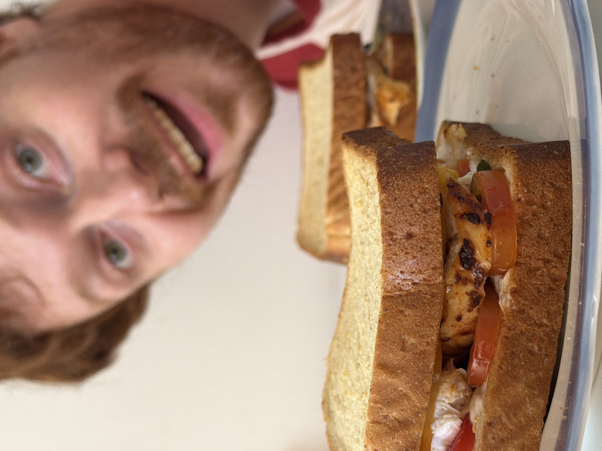 The scene is an indoor environment in Tel-Aviv, Israel. In the foreground, a sandwich, sliced diagonally, is presented on a white plate with a blue rim. The sandwich comprises two slices of white bread encasing grilled chicken portions exhibiting some char marks, red tomato slices, and a white creamy substance. A second, partially visible sandwich is situated in the mid-ground on a separate white plate. Occupying the upper right background, an out-of-focus adult male is depicted. He possesses reddish-brown hair and beard, light-colored eyes, and displays a broad smile, revealing teeth. A red and white garment is visible on his torso. The background consists of a plain, light-toned surface.