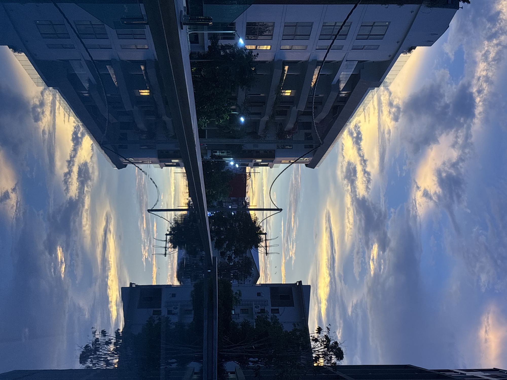The image shows a reflection of buildings and a sky with clouds in a window or mirrored surface. The location appears to be Choeng Thale, Thailand, based on the style of the buildings. The reflection creates a symmetrical image, with the buildings appearing both upright and inverted. The sky exhibits a blue hue with clouds that are highlighted by the setting or rising sun. Streetlights are also visible among the trees that surround the buildings. The buildings themselves are of a modern design, likely a residential or commercial complex.
