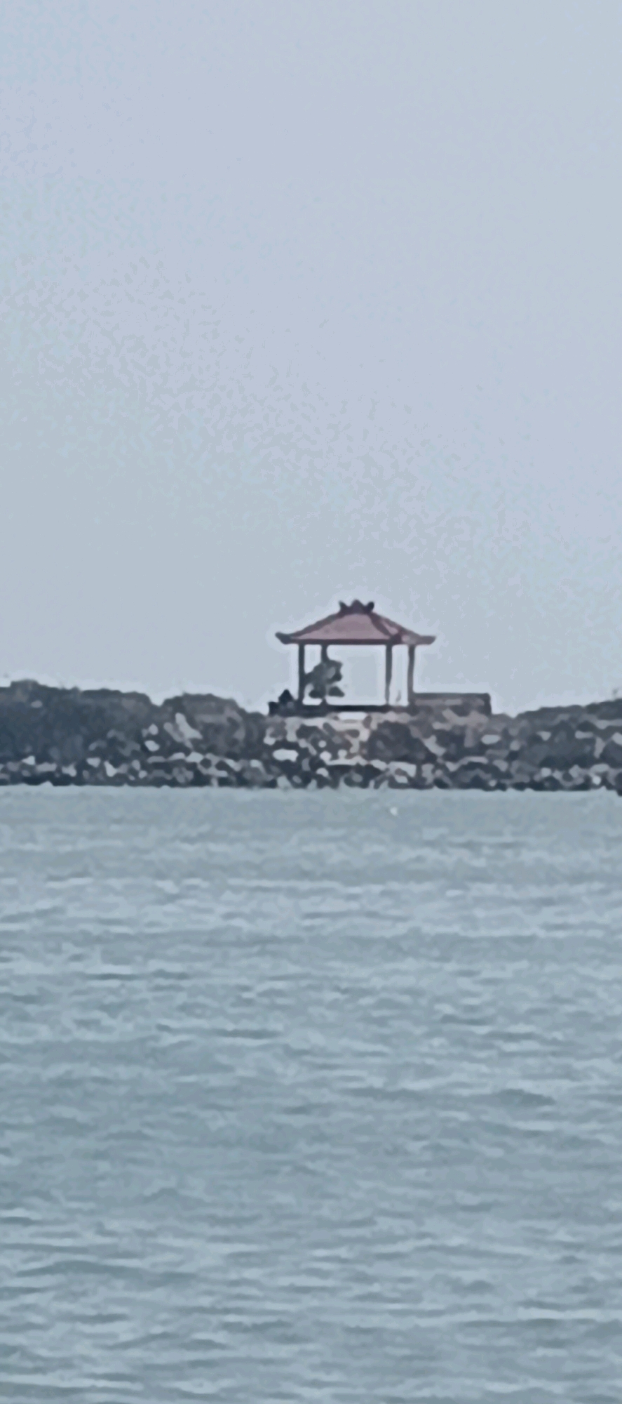 The image shows a small, red-roofed gazebo situated on a rocky outcrop overlooking a body of water. The gazebo has traditional architectural elements and appears to be empty. The water is calm with gentle ripples, and the sky is overcast. The rocky outcrop is covered with vegetation and appears to be a man-made structure, possibly a breakwater or part of a pier. The overall scene suggests a peaceful and somewhat isolated location. Given the information about Blimbing, Indonesia, this location could be a coastal area or a large lake within or near the city.
