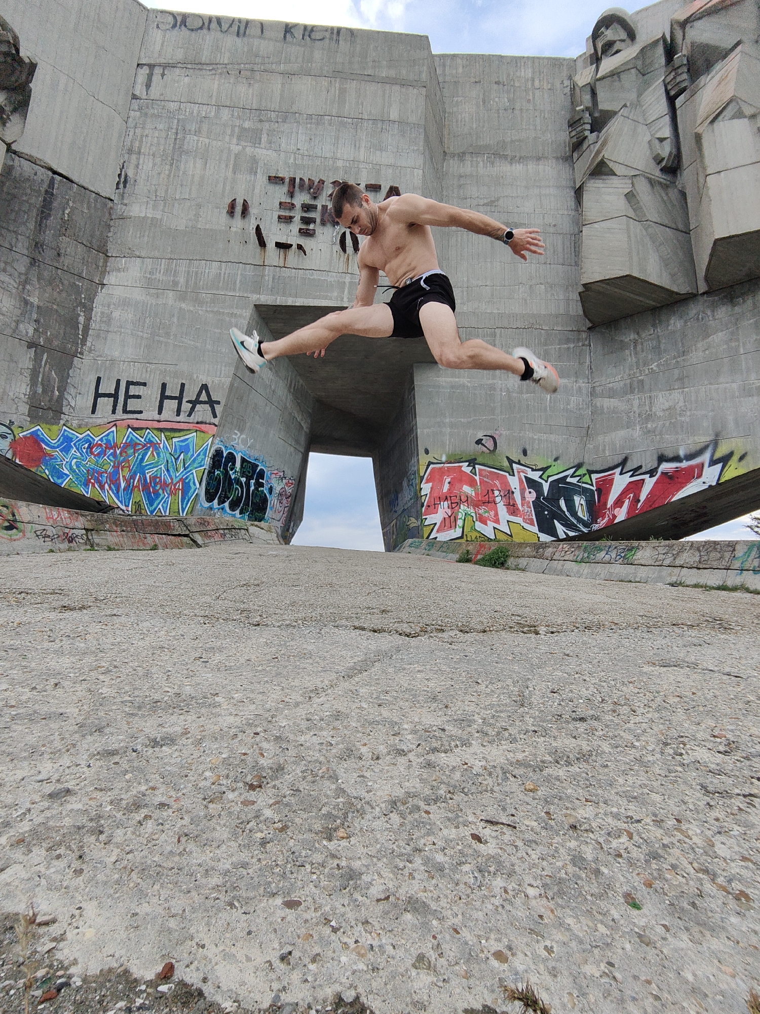 A man is suspended mid-air in a dynamic leap in front of a large concrete monument in Varna, Bulgaria. He has a muscular build and is wearing black shorts and sneakers. He is shirtless and his arms are outstretched. The monument behind him is a weathered, gray concrete structure, with graffiti art in vibrant colors on the lower sections. There is a visible archway through the monument, revealing the sky beyond. The ground is a rough concrete surface. The monument has inscriptions on the side and a sculpted relief on the right.