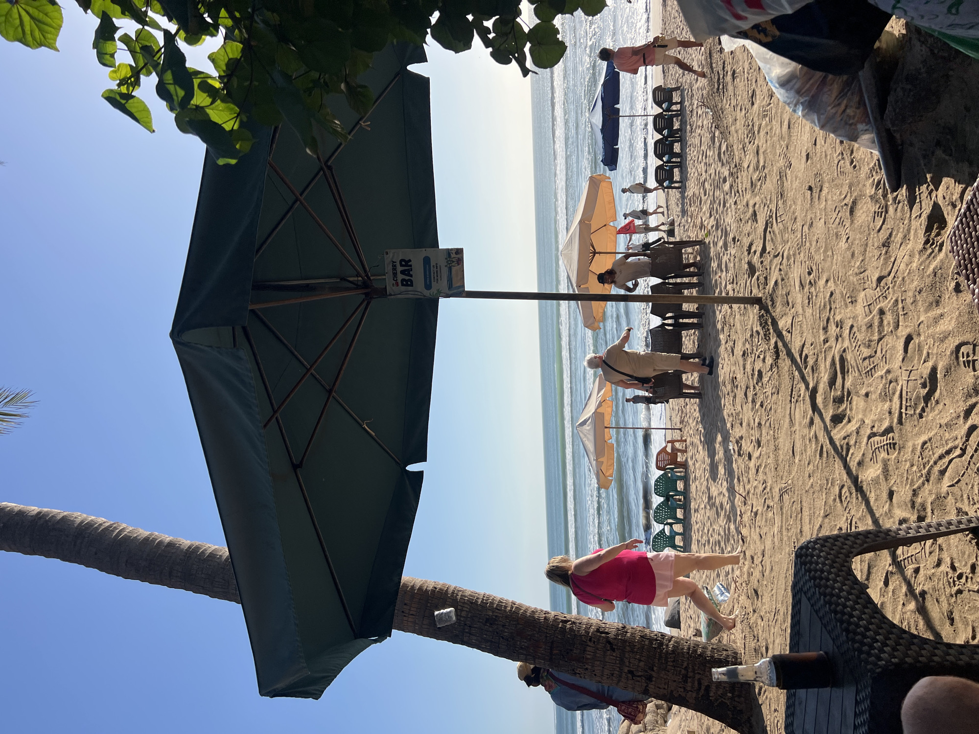 A wide-angle, eye-level outdoor shot captures a vibrant beach scene in Kuta, Indonesia, on a clear, sunny day. The foreground is dominated by a dark, wicker-style table with a bottle of brown liquid, presumably beer, resting on it. The sand is imprinted with numerous footprints, indicating recent activity.
In the mid-ground, a large, dark green beach umbrella provides shade. A sign attached to its pole reads "CHERRY BAR" with additional text below. Several beach chairs are arranged on the sand, some empty and some occupied by people. To the left, a tall palm tree stands prominently. A woman in a bright pink top and light-colored skirt walks away from the camera towards the ocean.
Further into the mid-ground, two lighter-colored beach umbrellas are set up. People are seen strolling along the shoreline, with the ocean and waves forming the backdrop. The water is a deep blue, with white crests indicating gentle waves. In the distance, more beach umbrellas and a few people are visible. To the far right, a person in a light-colored shirt and shorts walks towards the ocean.
The background consists of a clear blue sky, devoid of clouds, and the vast expanse of the ocean stretching to the horizon. The overall impression is one of a relaxed beach atmosphere, likely a popular spot for tourists in Kuta.