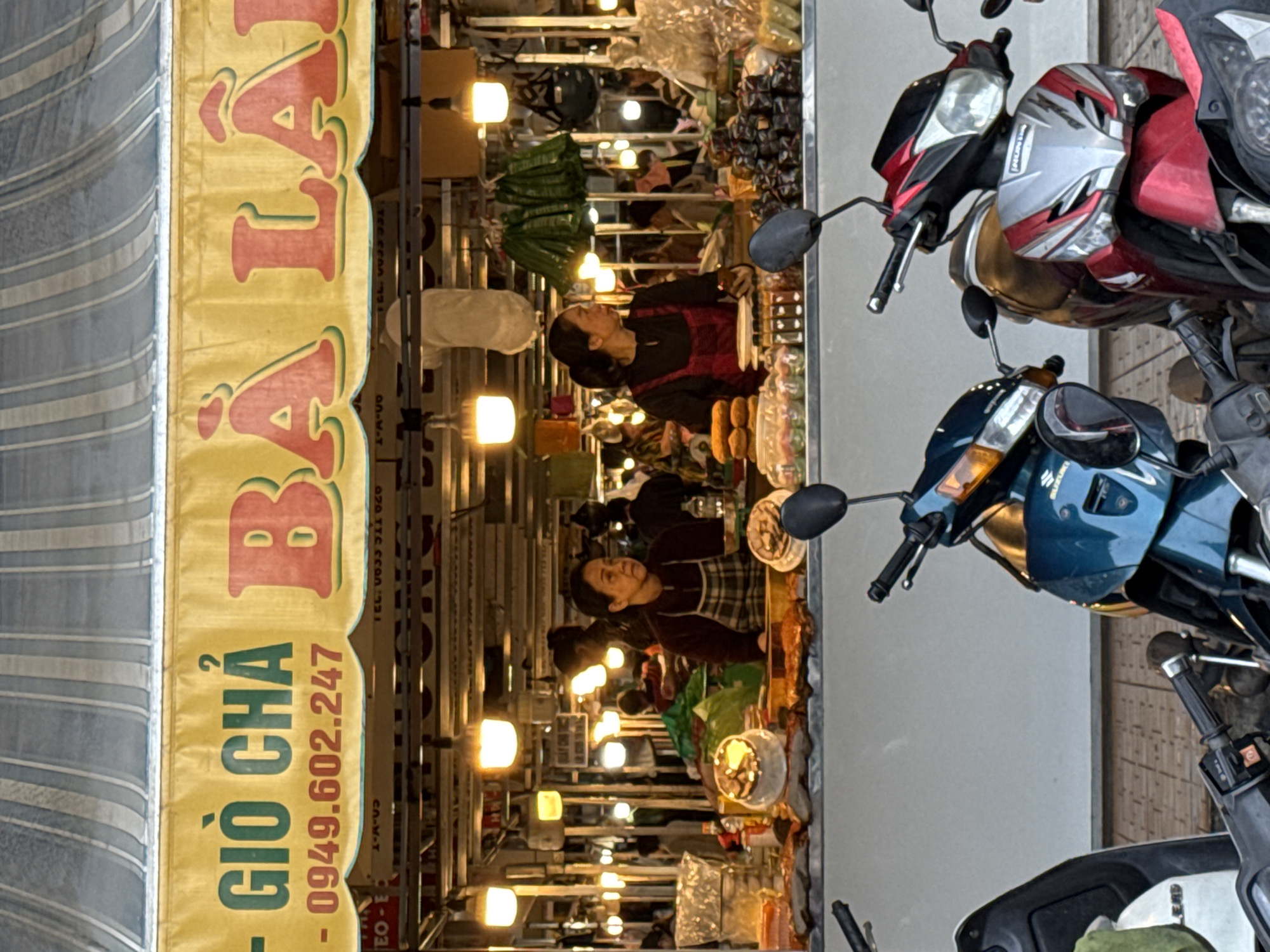 This is a medium shot, eye-level photograph taken from across a street, looking towards a food stall in Da Lat, Vietnam. The food stall is set up under a striped awning, with a large yellow banner above it displaying text in Vietnamese and a phone number. The text appears to be an advertisement for the food being sold, possibly "GIÒ CHẢ BÀ LÀ". Below the banner, the stall is illuminated by several hanging light bulbs.
Two women are visible behind the counter of the food stall. The woman on the left is wearing a dark top and a plaid apron, and appears to be looking towards her right. The woman on the right is wearing a dark top and a red apron, and is looking forward. Both women seem to be serving or preparing food.
The stall displays a variety of food items, including what appear to be meats, pastries, and bottled goods. In the foreground, several motorcycles are parked, partially obscuring the view of the stall. The ground in front of the stall is paved with interlocking stones. The lighting suggests it is either dusk or an indoor market with artificial lighting. The overall impression is that of a bustling street-side food vendor.