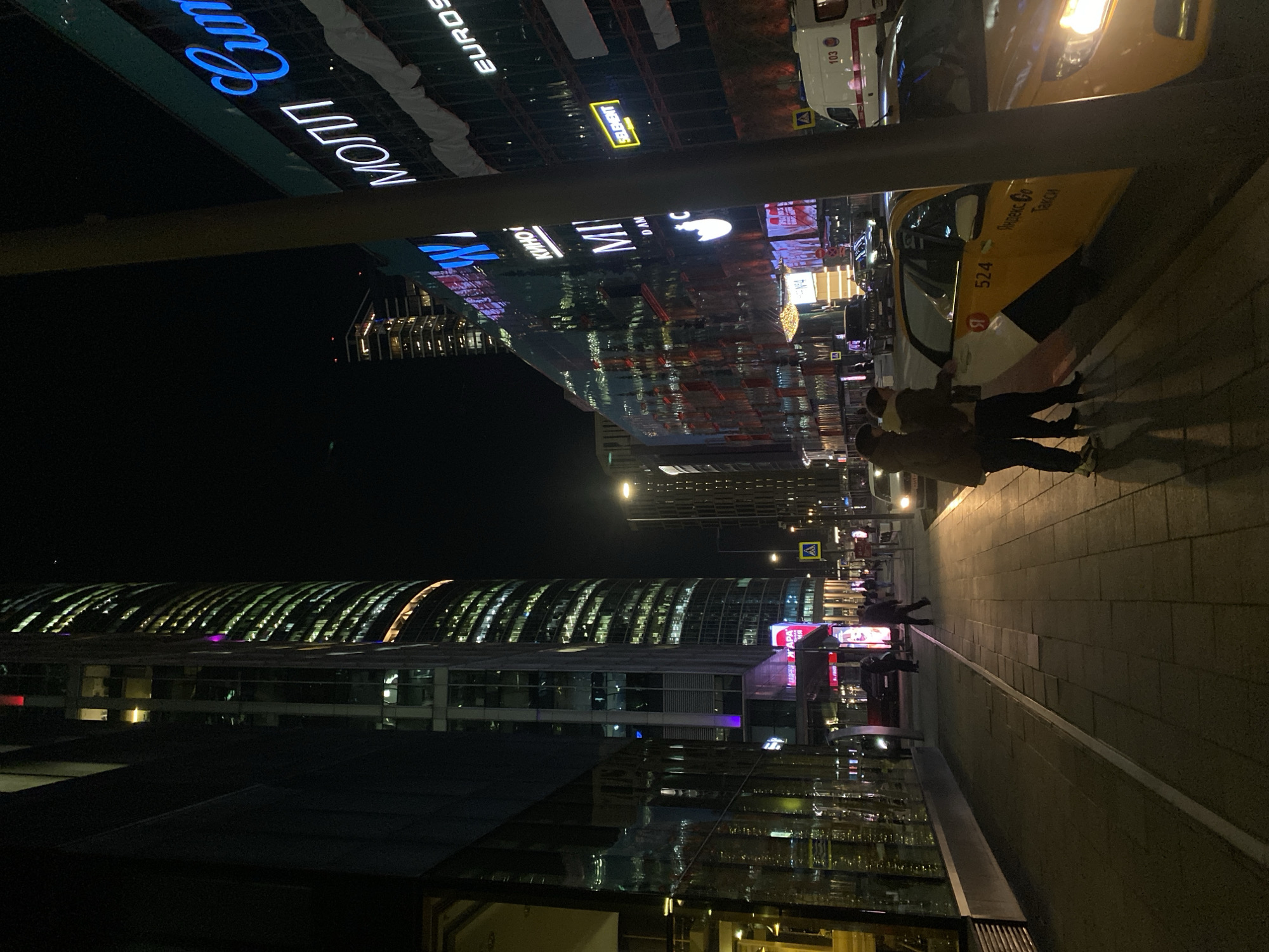 The image captures a nighttime scene in Khimki, Russia. In the foreground, two individuals are walking along a sidewalk next to a taxi. The sidewalk is paved with rectangular stones. The taxi, predominantly yellow and white, features the text "Яндекс Go Taxi" in Russian. An ambulance is parked further up the street. Above the taxi and ambulance, brightly lit commercial signs are visible, including recognizable brands like "EUROSET." In the background are several high-rise buildings with numerous illuminated windows. The reflections on the sidewalk are visible due to the damp or wet surface. The scene is illuminated by streetlights and lights from the nearby buildings.