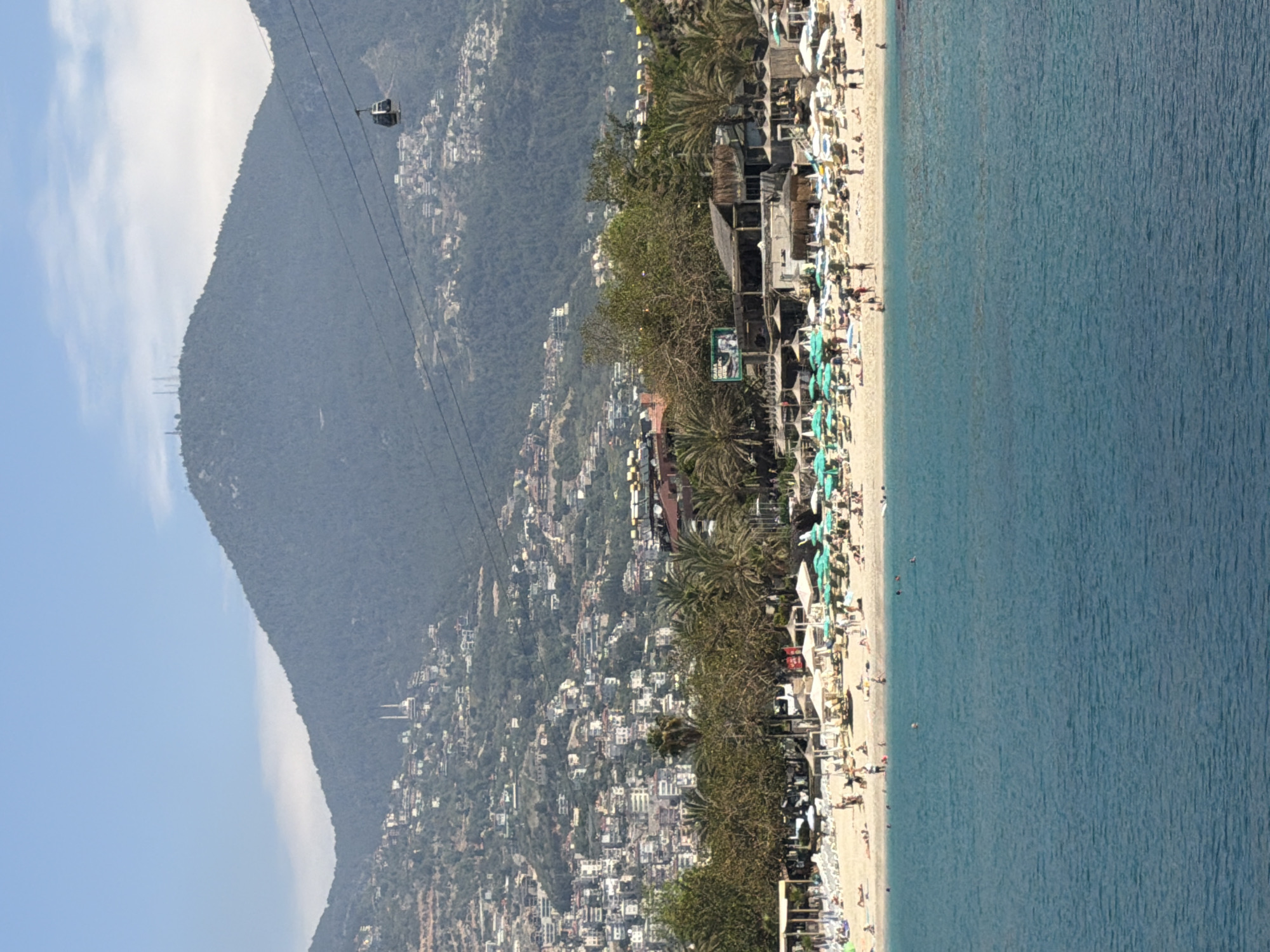 This image captures a vibrant coastal landscape encompassing a mountain, a bustling city, a sandy beach, and the clear blue sea, indicative of a popular holiday destination like Alanya, Turkey.
**Subjects:**
A large, green-covered mountain dominates the left and central background, its slopes densely populated with numerous white and light-colored buildings. A single cable car gondola is visible, traversing the mountain on dark suspension cables. Below the mountain, the city extends towards the coastline. The foreground features a long, sandy beach lined with an array of sun loungers, many of which are white, and umbrellas in shades of green and white/cream. Several wooden beach structures, possibly cafes or bars, are nestled among palm trees along the beachfront. The turquoise-blue sea occupies the right side of the image. The sky is bright blue with a few scattered white clouds.
**Activity or event taking place:**
The scene depicts typical leisure activities. People are visible on the beach, sunbathing and relaxing on loungers. Others are swimming or wading in the clear sea. The presence of the cable car suggests visitors are also engaged in sightseeing or transport up the mountain.
**Notable details:**
The time of day is bright daylight, likely mid-morning or early afternoon, given the clear sky and strong illumination. The weather is sunny and fair, ideal for beach and outdoor activities. The combination of a large mountain descending to a coastline, a sprawling city, a well-equipped beach, and a cable car system aligns well with the provided location context of Alanya, Turkey. The overall atmosphere appears to be one of relaxation and holiday enjoyment.
**Visible text:**
On a green structure near the beach, a small green sign with white text is partially visible. Due to the distance, the text is not entirely clear but appears to read "CAFE B..." suggesting a beach cafe or bar.