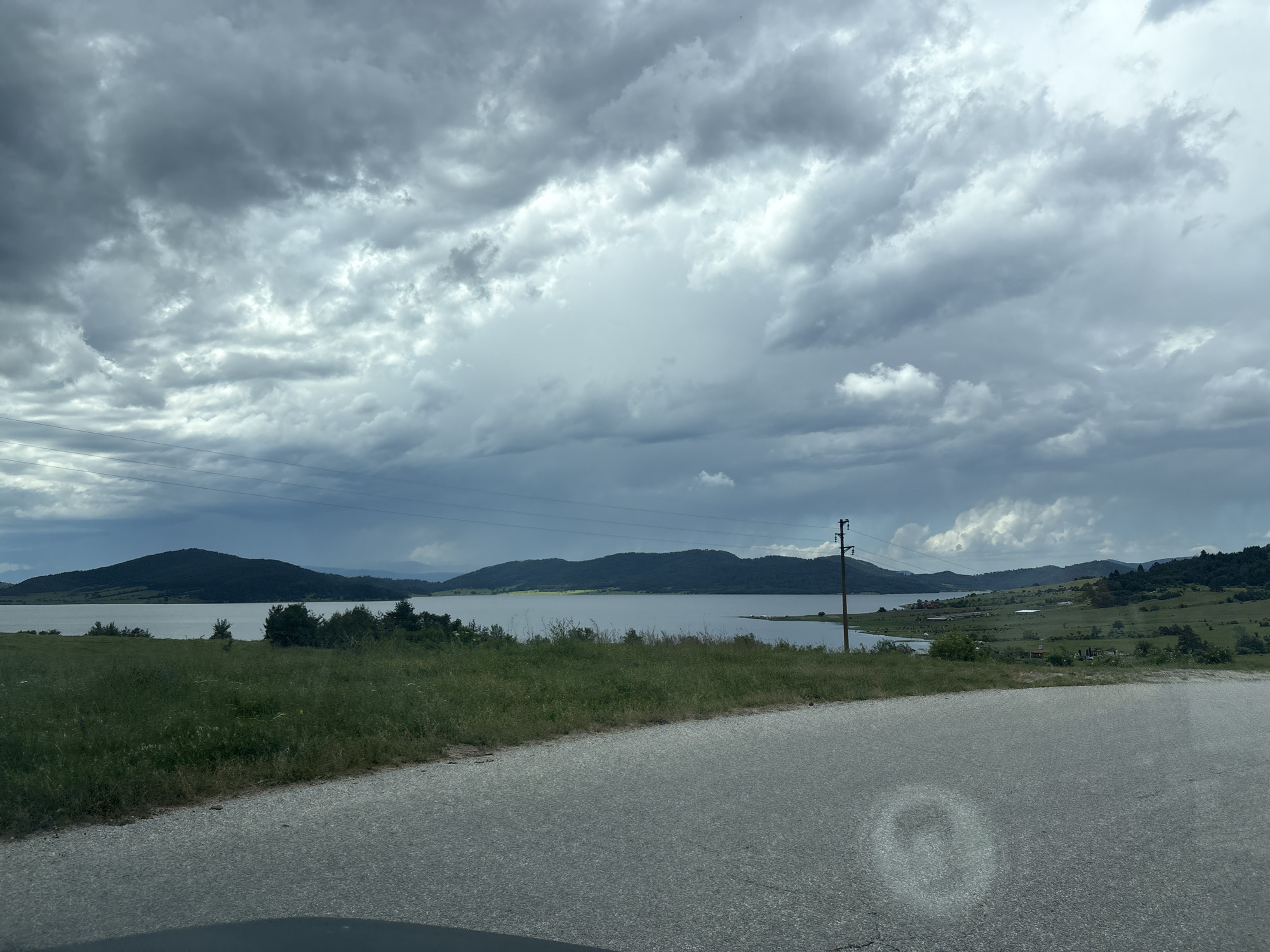 The image depicts a scenic view of a large lake, likely Batak Reservoir, in Batak, Bulgaria. The sky is overcast with dramatic, heavy clouds, suggesting impending rain or stormy weather. A paved road curves into the foreground from the right, with a grassy embankment bordering it on the left. Beyond the embankment lies a stretch of green vegetation leading to the tranquil waters of the lake. The far side of the lake is framed by a range of undulating, tree-covered hills and mountains. A single utility pole with electrical wires is visible on the right side of the frame, a subtle indication of human presence in the otherwise natural landscape. The overall mood is somber and dramatic, with the diffused light accentuating the textures of the clouds and the natural contours of the land.
