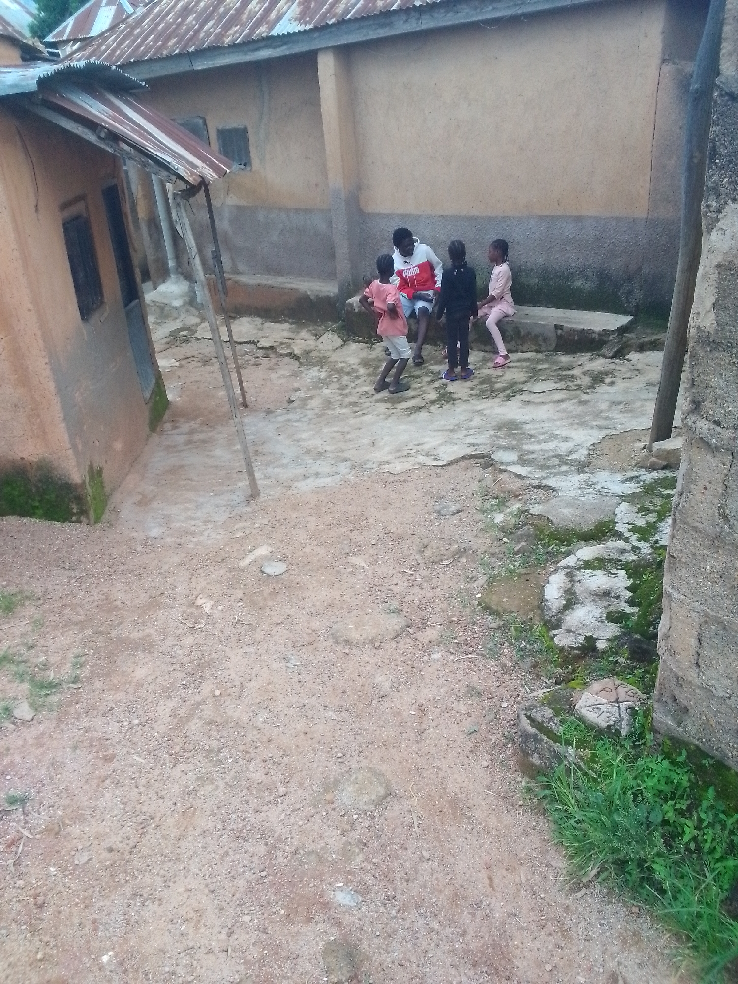 In an outdoor setting that appears to be a village or informal settlement, a group of four individuals, three children and one adult male, are gathered. The adult male is seated, wearing a red and white hooded jacket and blue shorts. He is facing left, interacting with the children. To his right, a young child wearing a pink shirt and white shorts stands. To his left, a young child in a black top and black leggings is standing, and another child, wearing a pink top and pink leggings, is seated.
The ground is a mixture of dirt, gravel, and concrete patches. Some areas show signs of moss and patchy grass growth, suggesting a humid environment. The background features two dilapidated structures with corrugated metal roofs and mud or concrete walls. One structure on the left has a sloping roof and visible wooden supports. The other structure on the right is partly obscured by a concrete pillar. The overall scene suggests a humble, rural or semi-urban environment.
