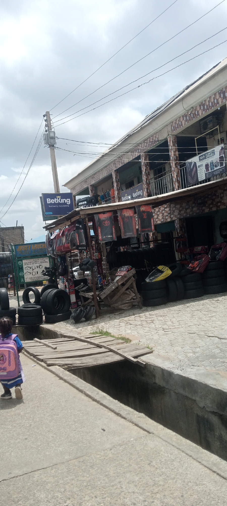 The image depicts a street scene in Jalingo, Nigeria, near the BetKing betting shop. The foreground shows a young child with a pink backpack walking on a paved walkway, which leads to a wooden plank bridge crossing a drainage ditch. Beyond the walkway, a variety of car tires are stacked on the ground. In the background, a building with multiple floors is visible, with shops on the ground level. One shop features a BetKing sign, and displays of car accessories, including what appear to be car floor mats with logos like "TRD" and "TOYOTA," hang from the storefront. Several individuals are standing on the balcony of the building. The sky is overcast. The overall scene suggests a commercial area with businesses catering to vehicle owners and offering betting services.