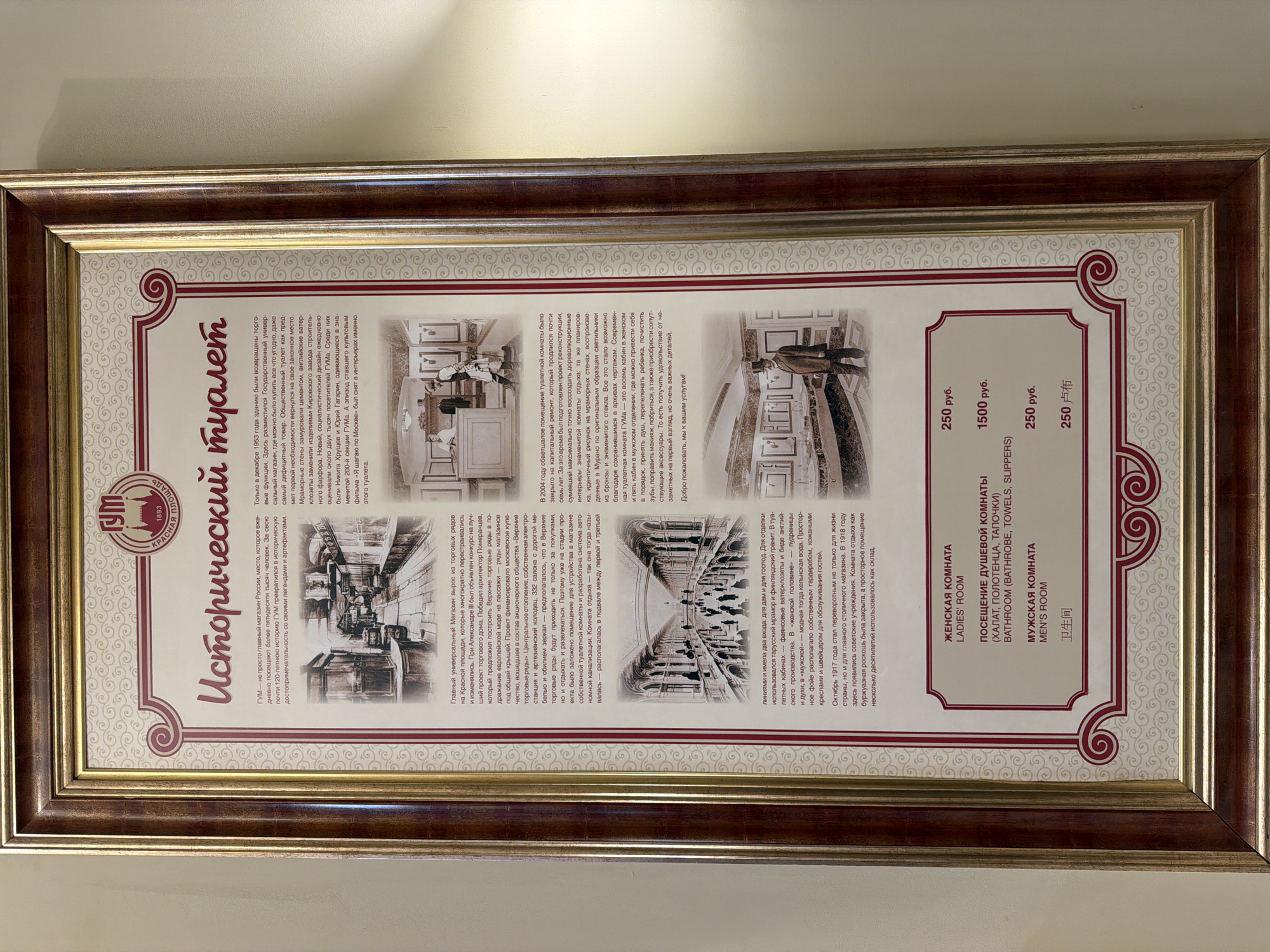 The image shows a framed informational sign about the historical toilet facilities at the GUM department store on Red Square in Moscow, Russia. The sign includes a title in Russian, "Исторический туалет" ("Historical Toilet"), along with photographs of the toilet facilities in the past and present. Text provides details on the history of the toilet facilities, from their construction in the late 19th century to their renovations and modern amenities. Pricing is provided in Rubles (руб) for access to the various facilities, including the ladies room, bathroom (with bathrobe, towels, slippers), and men's room.