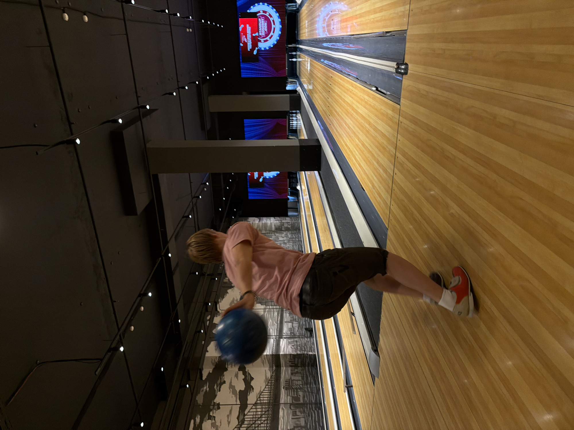 In an indoor bowling alley, a person with blonde hair, wearing a pink t-shirt and dark shorts, is in the process of throwing a blue bowling ball down a lane. The lane is made of polished wood with inlaid strips, and the background shows more lanes, digital screens displaying graphics, and a mural of a city skyline with a bridge. The ceiling is dark with numerous track lights. The person is mid-action, with their arm extended, and their body angled to propel the ball. This bowling alley is located in the city of Izhevsk, Russia.