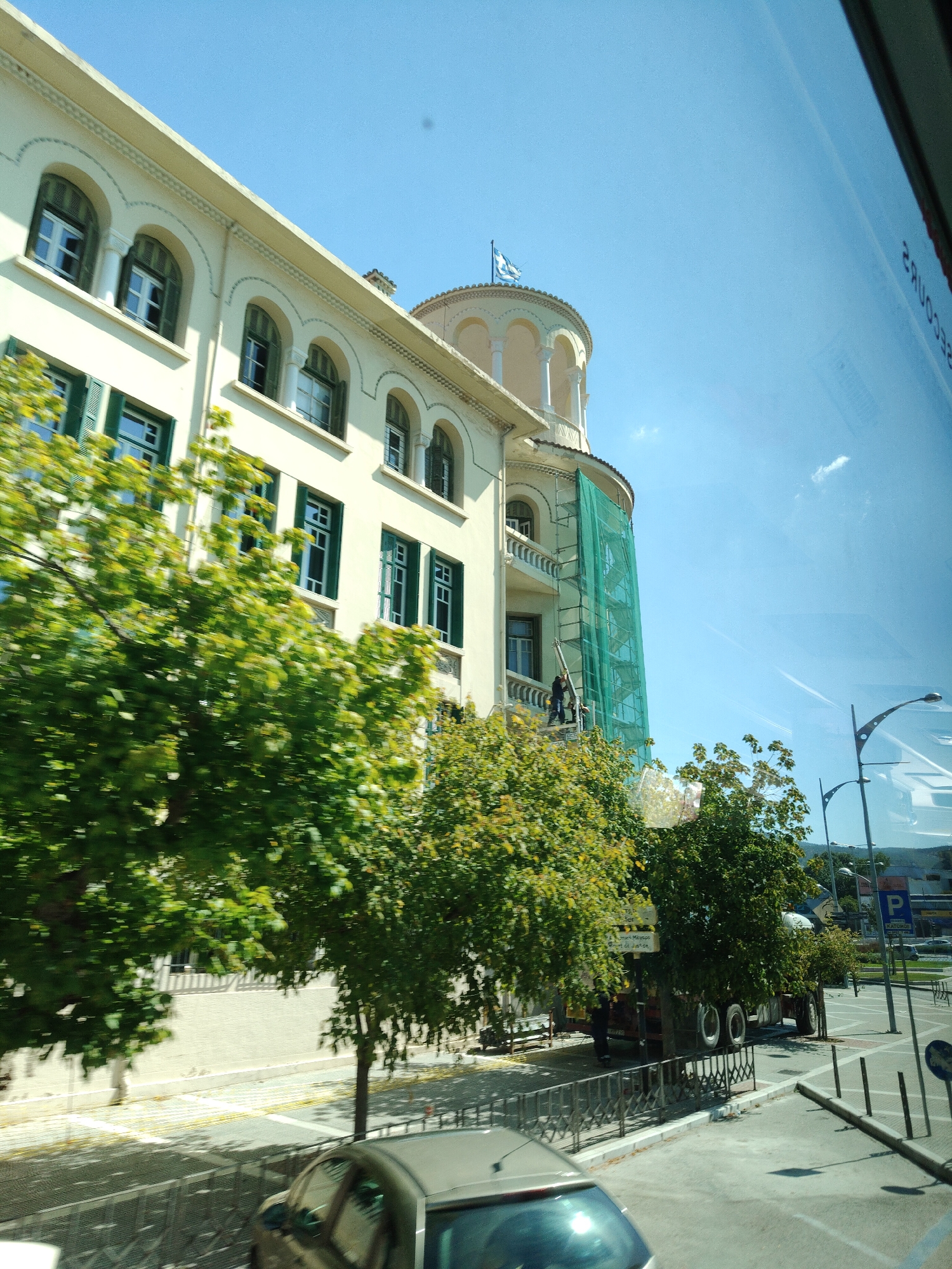 The image captures a sunny day in Thessaloniki, Greece, showcasing a building undergoing renovation. The building, a multi-story structure with a light-colored facade, features arched windows with dark green shutters. Scaffolding covered in green netting is erected on the side of the building, indicating ongoing construction work. Two individuals are visible on the scaffolding, appearing to be engaged in their tasks.
In the foreground, lush green trees with dense foliage partially obscure the building, suggesting a street-side view, likely from a vehicle. A silver car is visible in the lower part of the frame, its reflection caught on the surface, hinting at a reflection from a window. Beyond the trees, a truck with two visible wheels is parked. Further in the background, streetlights and a blue sign with the letter "P" are noticeable, suggesting an urban environment with parking facilities. The clear blue sky overhead, with a few faint clouds, suggests pleasant weather.
