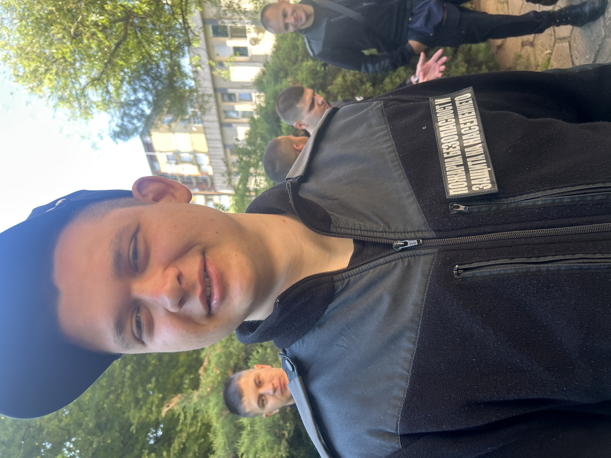 A young man wearing a black baseball cap and a black jacket with a patch that reads "ПОЖАРНА БЕЗОПАСНОСТ И ЗАЩИТА НА НАСЕЛЕНИЕТО" is facing the camera. Several other men are visible in the background, some in similar attire. The location appears to be outdoors in an urban environment, possibly in Sofia, Bulgaria. There is greenery in the background, along with a building.