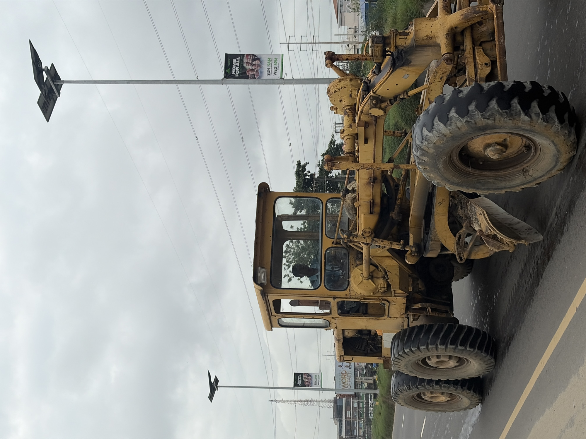 A yellow motor grader is driving on a road in Alaguntan, Nigeria. A man is operating the grader from inside its cab. The sky is overcast. In the background, there are power lines, streetlights, and buildings. A banner on a streetlight reads "HOME FOR ALL" and lists service times. Another banner in the distance is partially obscured, but appears to be related to a church or religious organization. The road surface is asphalt with yellow lane markings. The grader has large, rugged tires, suggesting it is being used for construction or road maintenance.