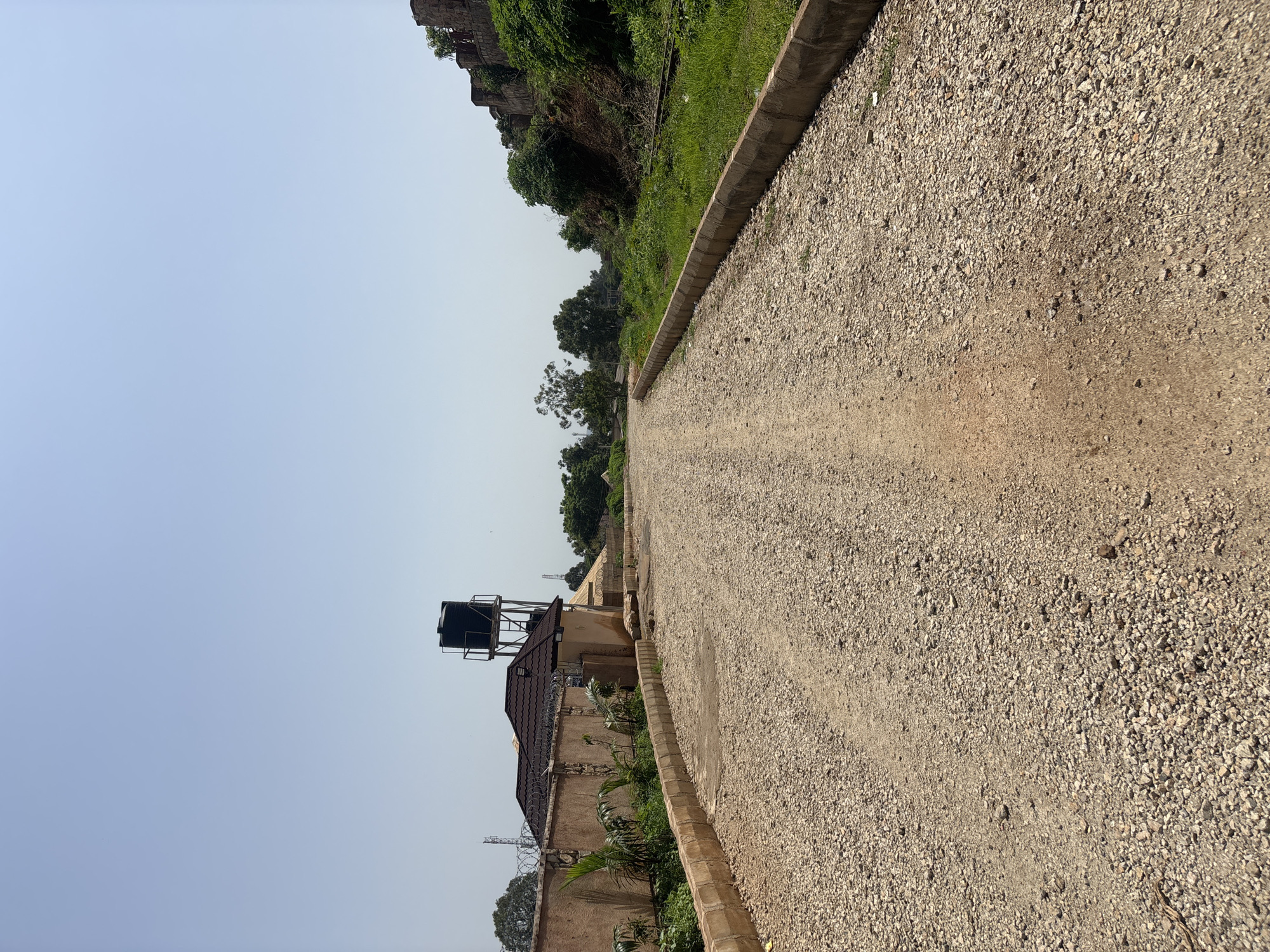 The image depicts a wide, unpaved gravel road curving into the distance under a clear, pale blue sky. The road is flanked by concrete curbs on both sides. To the left, a textured tan wall with barbed wire fencing encloses a property with a house featuring a dark, multi-gabled roof and a tall black water tank on a metal structure. Lush green vegetation, including palm-like plants, grows along the inside of the wall. In the distance on the left, telecommunication towers are visible. To the right of the road, a grassy slope rises, topped by a weathered stone wall or fortification, with dense green foliage and trees growing along its base and top. The overall scene suggests an undeveloped or under-construction area within a city, possibly on the outskirts, given the mix of urban infrastructure (water tank, towers) and natural elements. The location is identified as the City of Jos, Nigeria, implied by the context of the query.