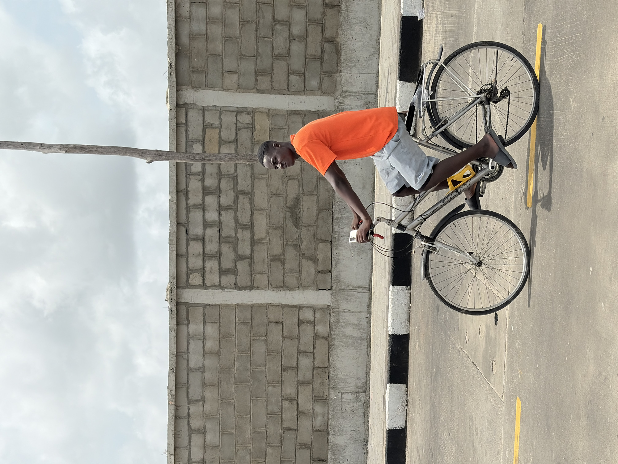 A young man rides a gray Giant bicycle on a paved road in Igboefon, Nigeria. He wears an orange t-shirt and gray shorts, along with dark-colored slide-on sandals. The road has a black and white painted curb on one side and a yellow painted line on the opposite side. A concrete block wall stands adjacent to the road. A thin, weathered wooden pole extends horizontally from the wall into the sky. The sky is partly cloudy.