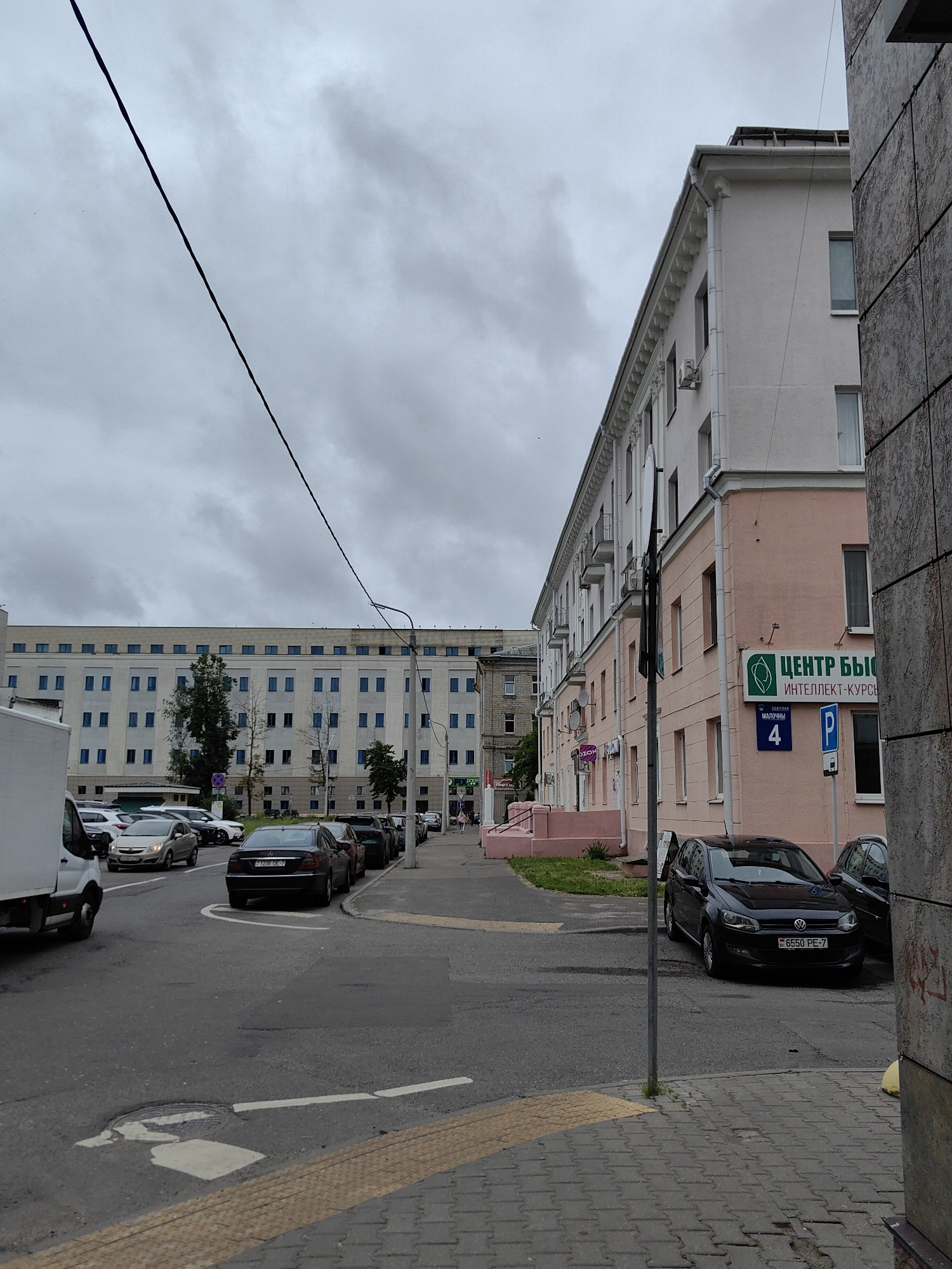 The image captures a street scene in Minsk, Belarus, with a focus on a row of buildings and parked cars under an overcast sky. On the right side of the frame, a light pink building with white trim and multiple windows is prominent. A green sign with white text reading "ЦЕНТР БЫСТ", followed by "ИНТЕЛЛЕКТ-КУРСЫ", is affixed to the building. Below this, a blue sign with a white 'P' indicates parking. A dark Volkswagen Polo is parked in front of this building. To the left, a long, light-colored building with a series of evenly spaced blue windows stretches into the distance. Several cars are parked along the street in front of both buildings. A white van is visible on the far left. Power lines crisscross the sky. The street is paved, and a sidewalk with textured yellow tactile paving is present in the foreground.