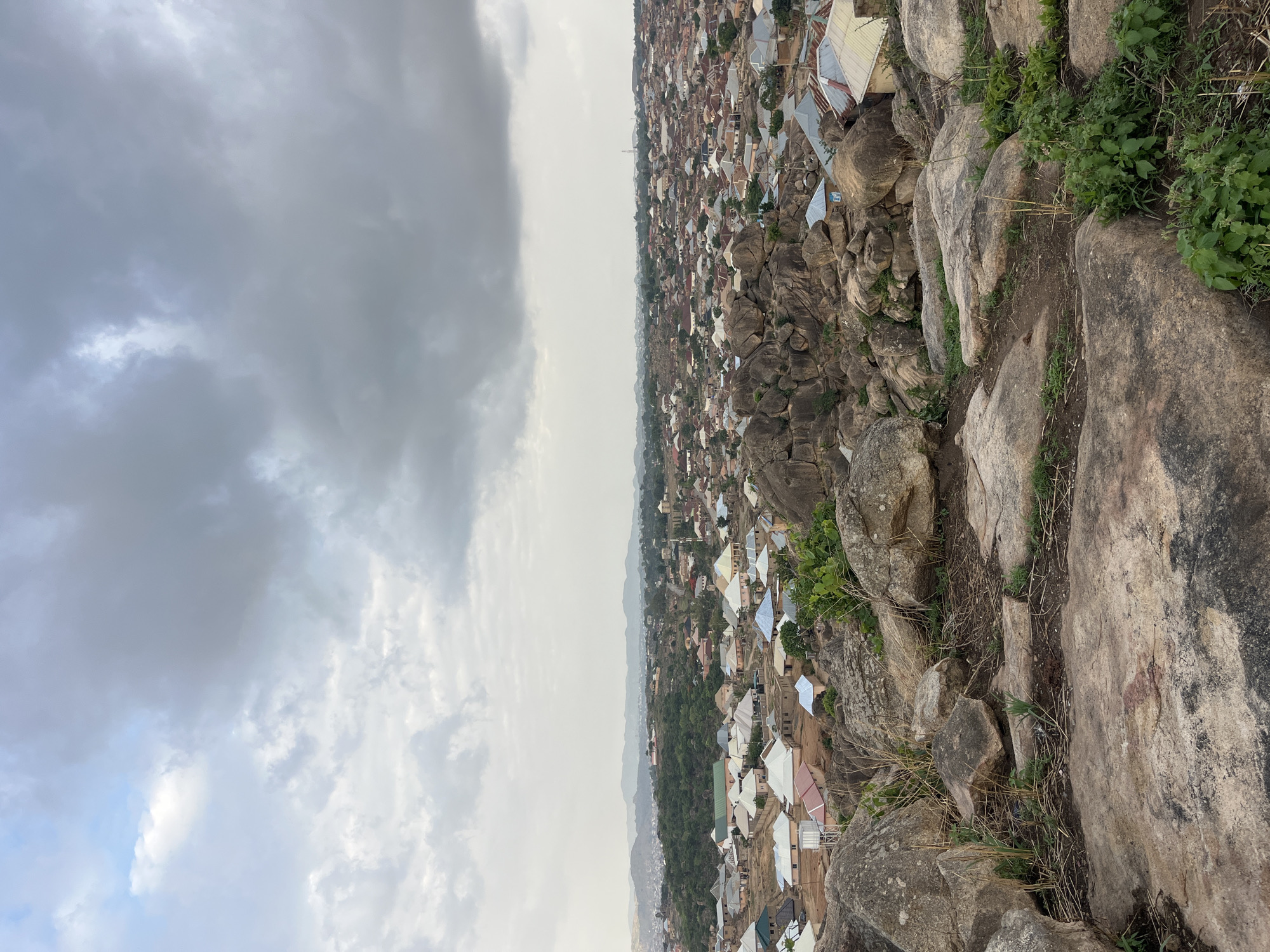 The image shows an elevated view of a section of Jos, Nigeria, from a rocky outcrop or hill. The foreground consists of large, weathered rocks and sparse vegetation. The middle ground shows a dense urban area with numerous buildings, many of which have metal roofs. In the background, the landscape fades into a hazy horizon with hills visible in the distance. The sky is overcast with heavy clouds.